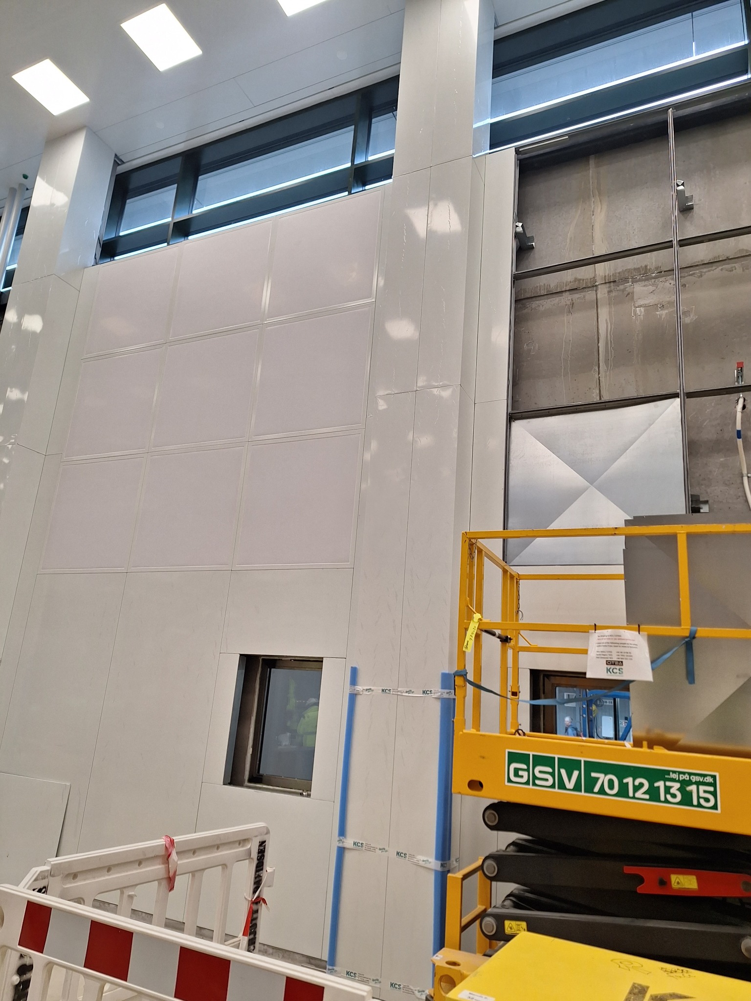 Montaż białych paneli ściennych w pomieszczeniu z oknami, widoczny podnośnik GSV i barierki. Panel z małym oknem. Częściowo widoczna surowa ściana.