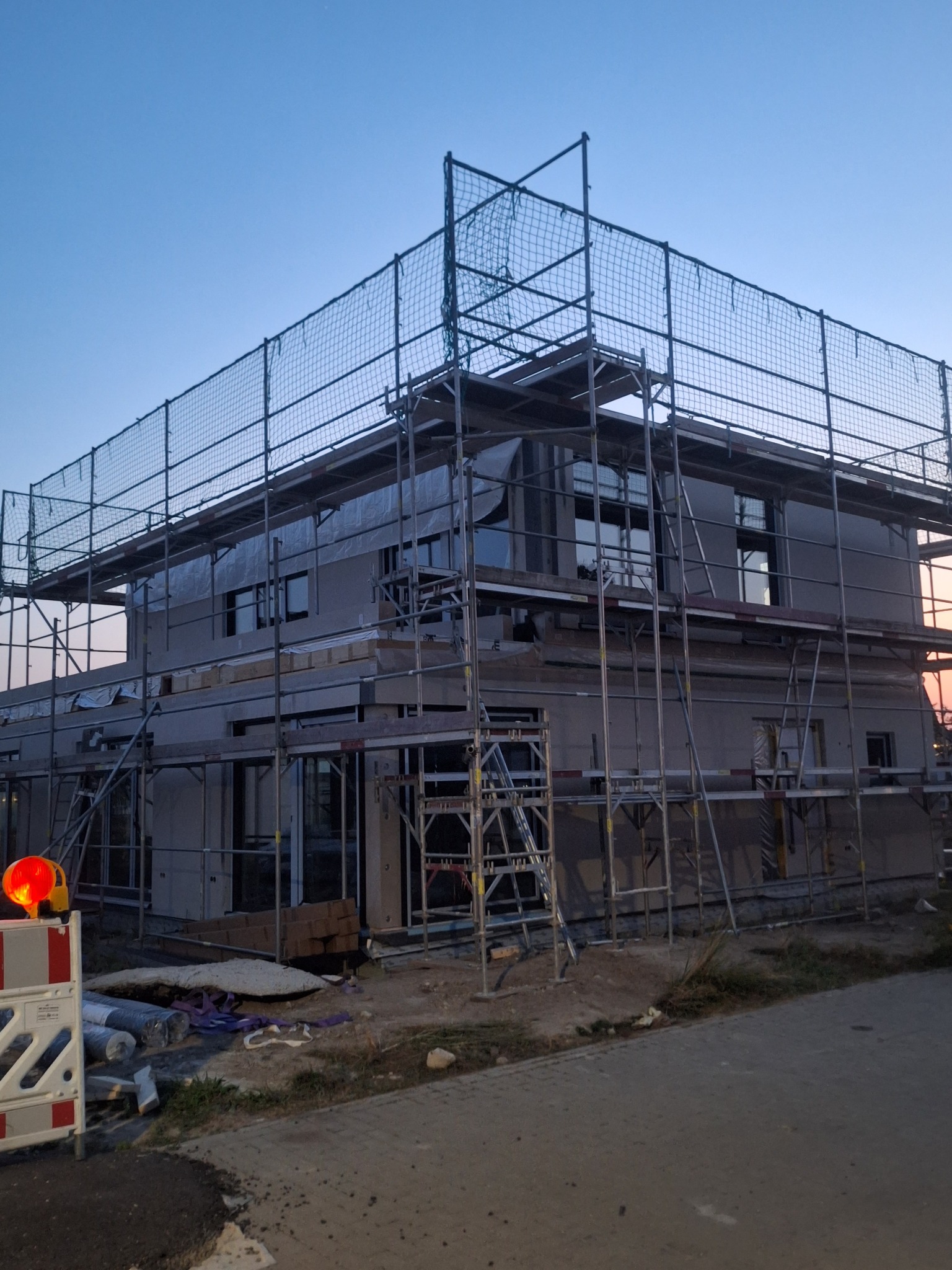 Dwukondygnacyjny dom w trakcie budowy, otoczony rusztowaniami i siatką zabezpieczającą, widoczny o zmierzchu. Elementy konstrukcyjne i materiały budowlane w tle.