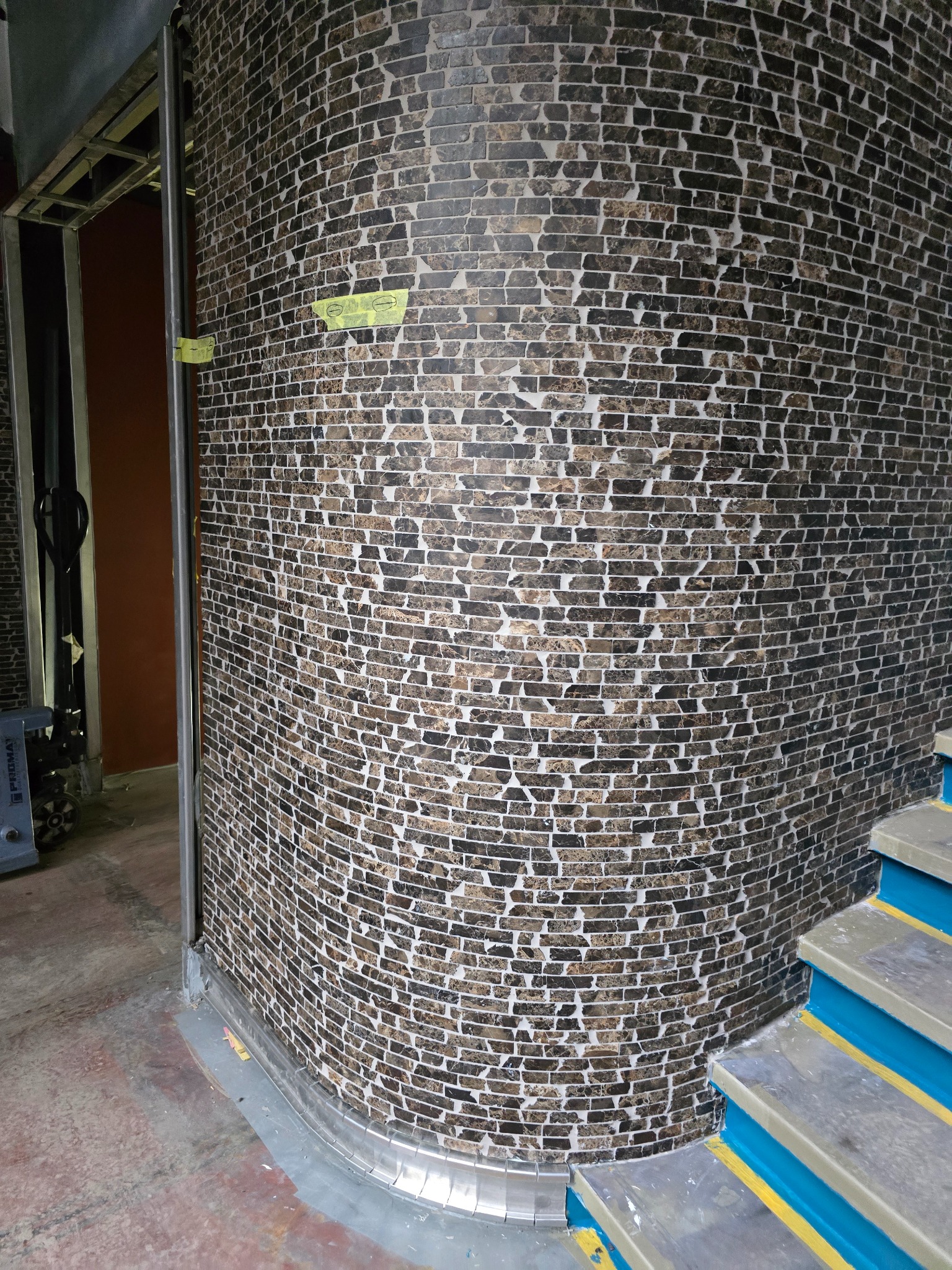 Wykończenie ściany mozaiką imitującą cegłę, widok na fragment schodów z niebieskimi akcentami. W tle metalowa konstrukcja i fragment podłogi.