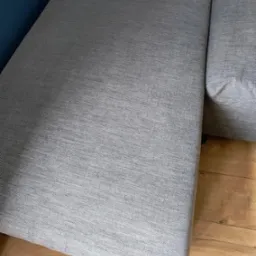 Szary narożnik z tkaniny o drobnej fakturze, widoczny z bliska, z fragmentem drewnianej podłogi i ciemnoniebieskiej ściany w tle. Dobre oświetlenie podkreśla detale materiału.