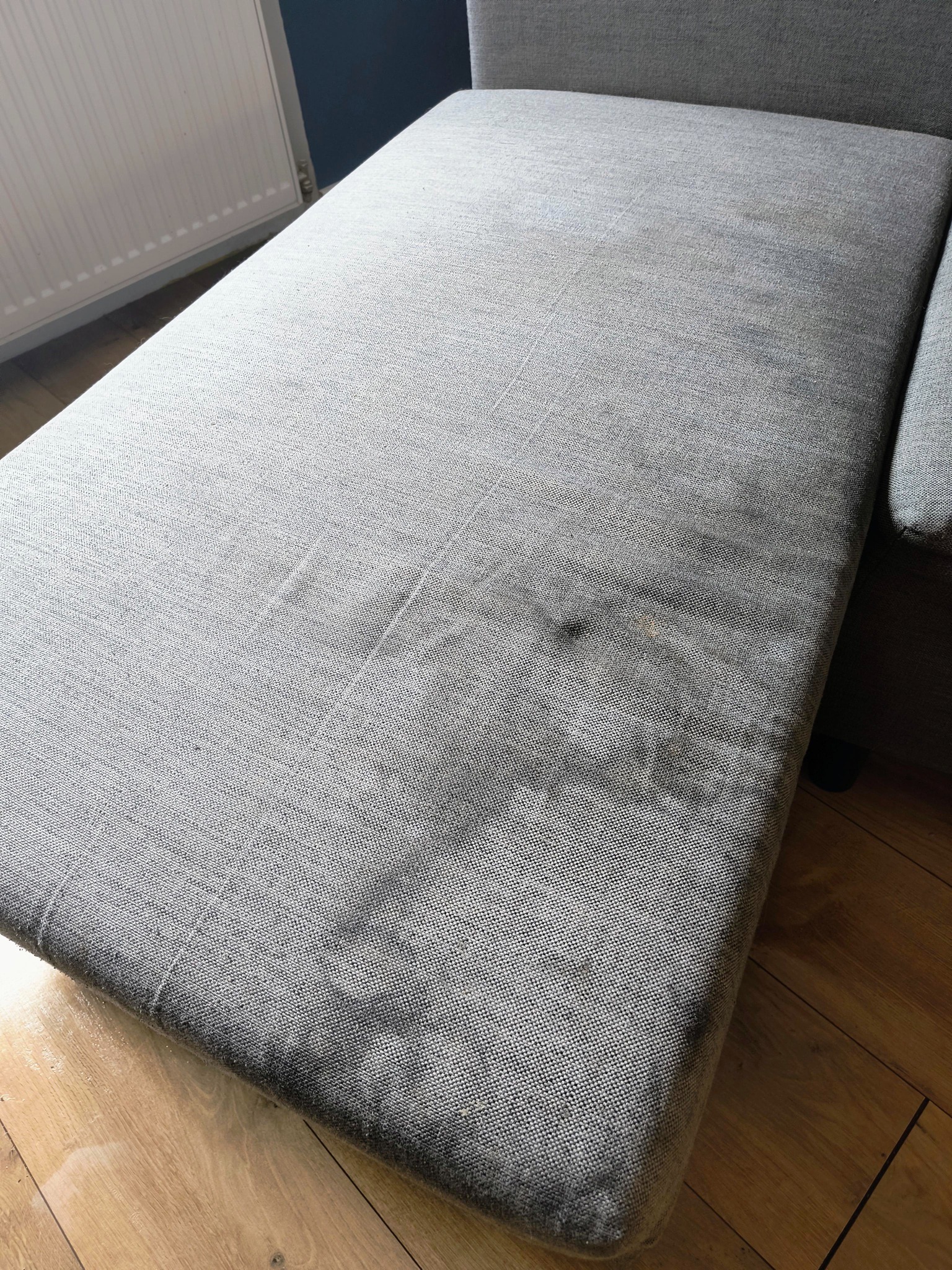 Zabrudzona, szara kanapa z plamami na tapicerce, widoczna na tle drewnianej podłogi w Bytomiu. Wymaga odświeżenia i profesjonalnego czyszczenia.