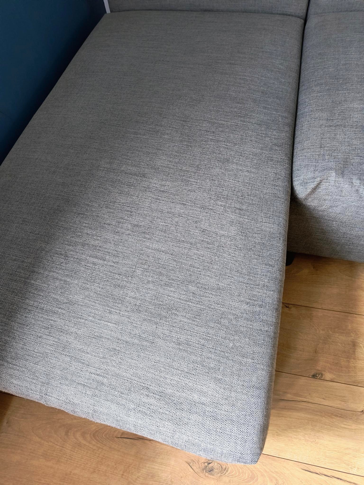 Szary narożnik z tkaniny o drobnej fakturze, widoczny z bliska, z fragmentem drewnianej podłogi i ciemnoniebieskiej ściany w tle. Dobre oświetlenie podkreśla detale materiału.