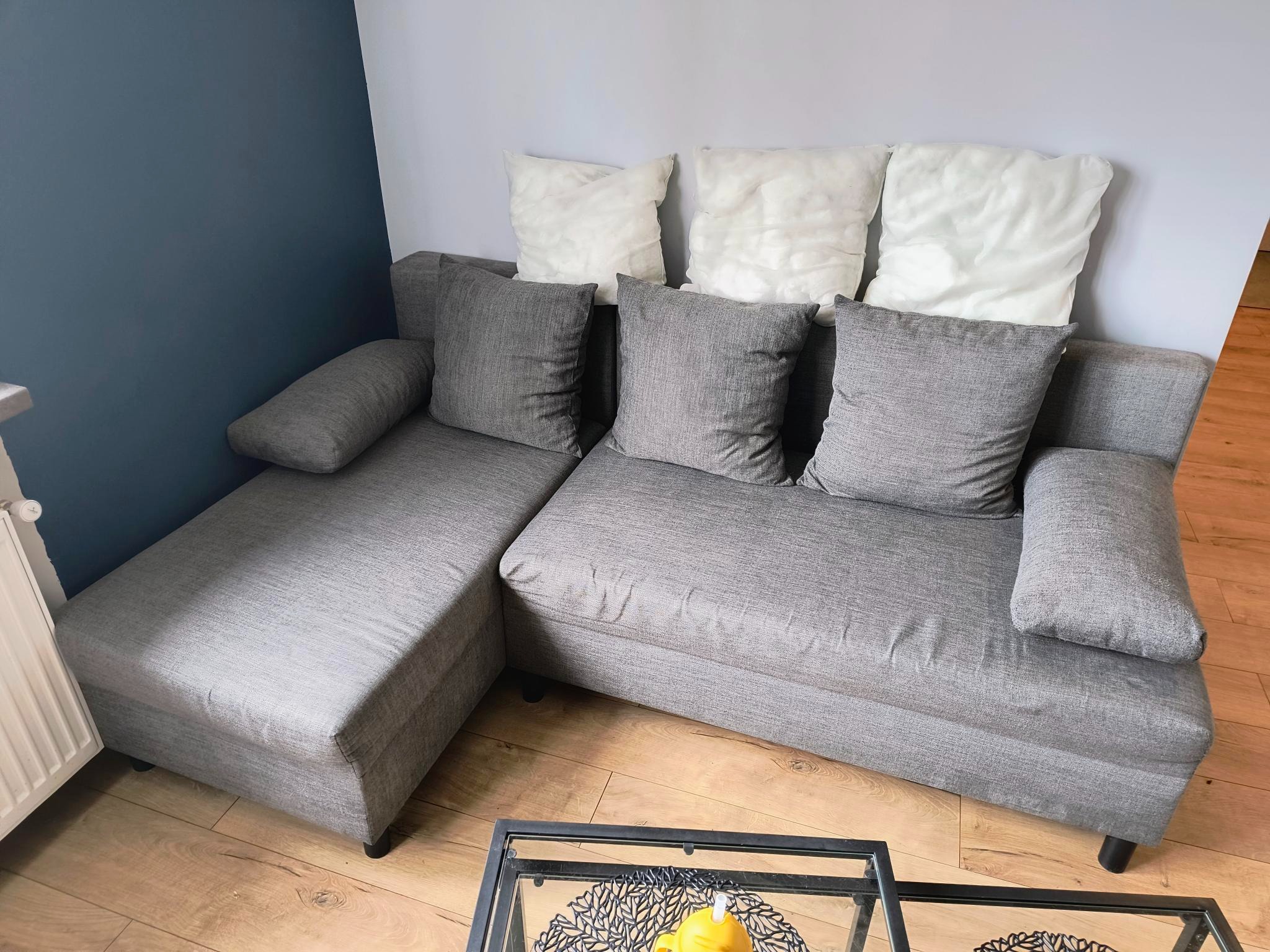 Szara, narożna kanapa z poduszkami na drewnianej podłodze, z widocznym stolikiem kawowym w tle. Neutralne oświetlenie.