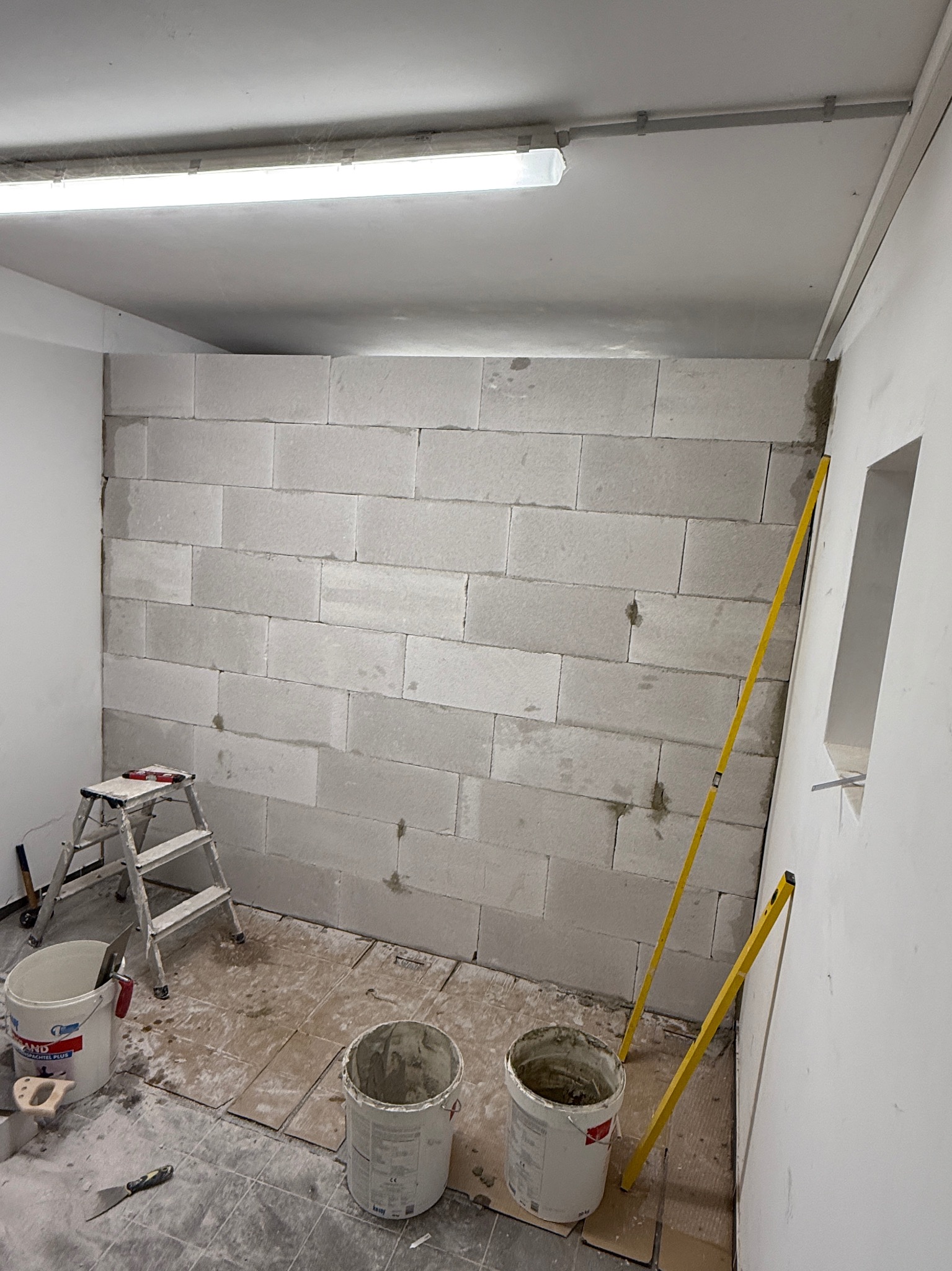 Wznoszenie ściany z bloczków betonu komórkowego wewnątrz pomieszczenia. Widoczne wiadro z zaprawą, poziomica i drabina. Praca w toku, surowy stan.