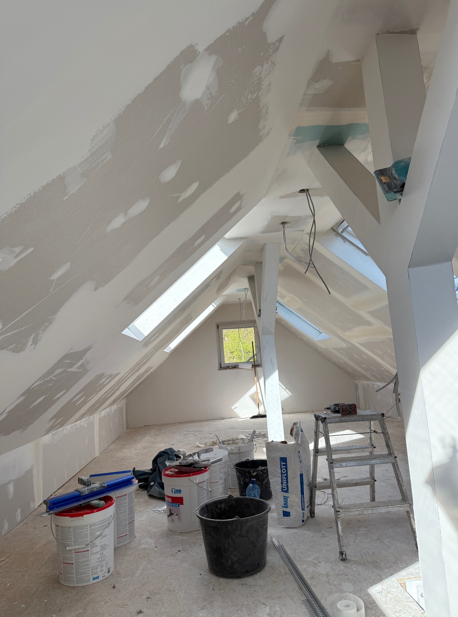 Poddasze w trakcie szpachlowania: ściany skośne z oknami dachowymi, widoczne narzędzia, drabina, worki z gładzią. W tle okno z widokiem na zieleń.