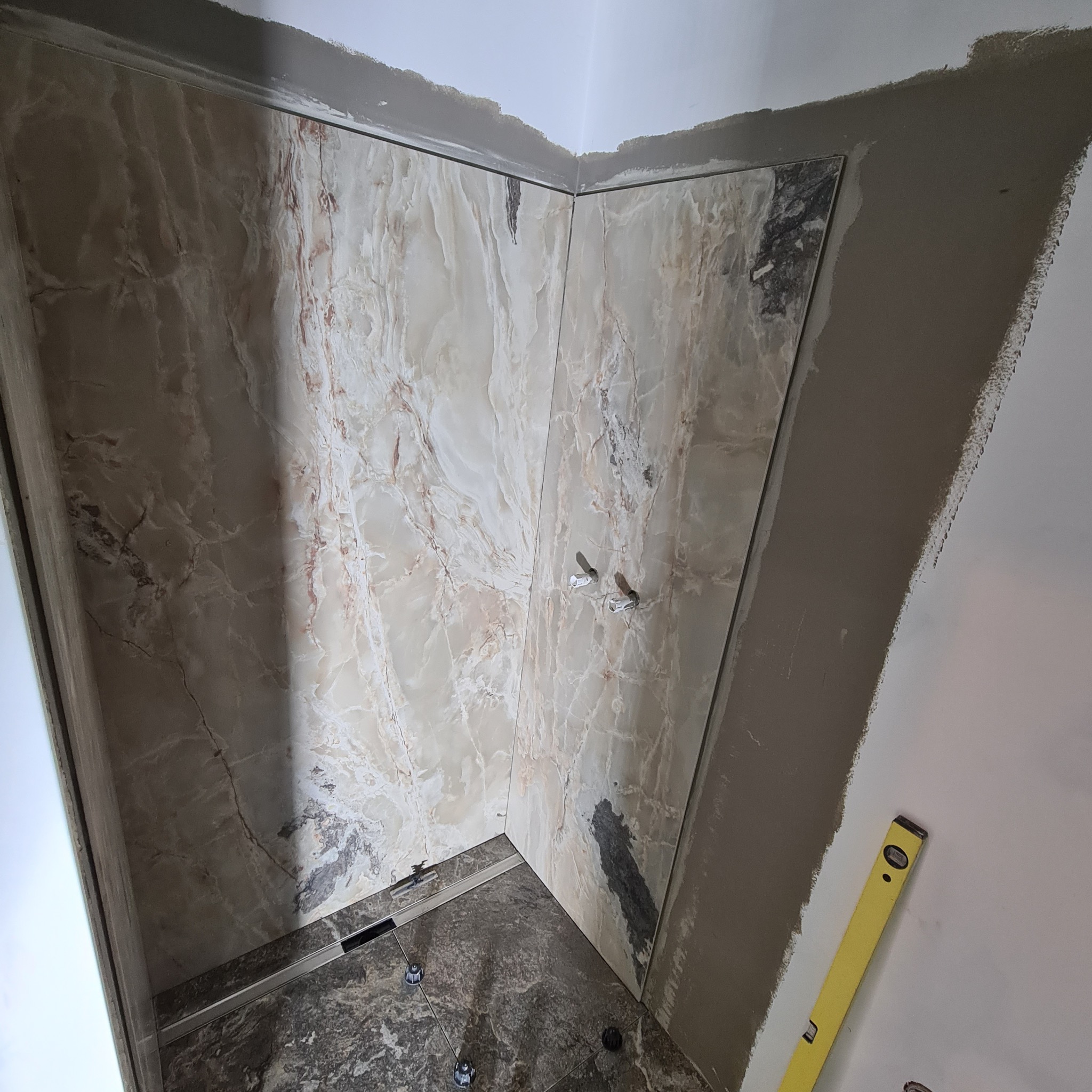 Narożnik łazienki w trakcie wykańczania: marmurowe płytki na ścianach, widoczne mocowania armatury, poziomica przy ścianie, świeżo nałożona zaprawa.