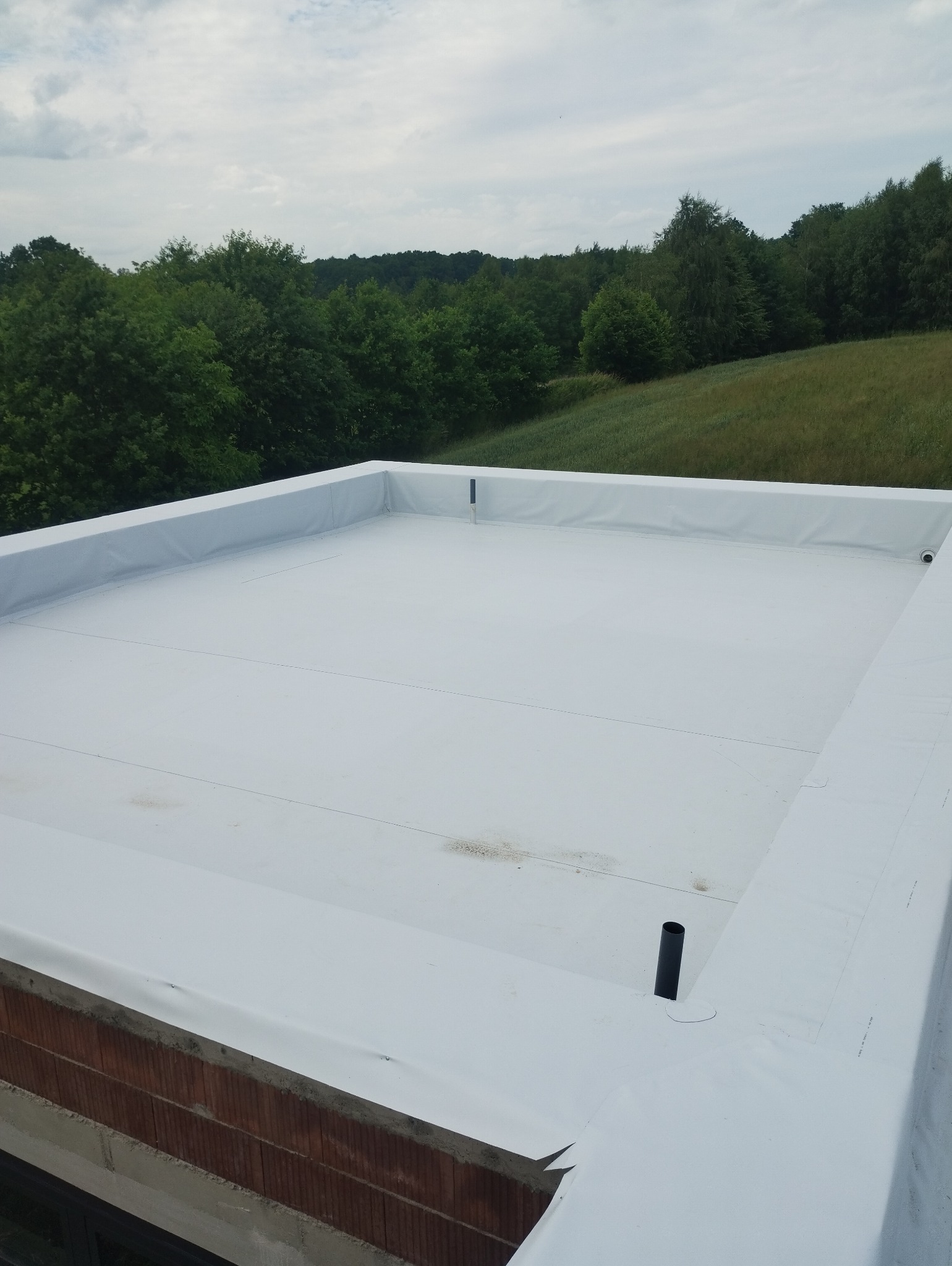 Biała membrana dachowa EPDM na płaskim dachu w trakcie montażu, widoczne detale mocowania i krajobraz w tle.