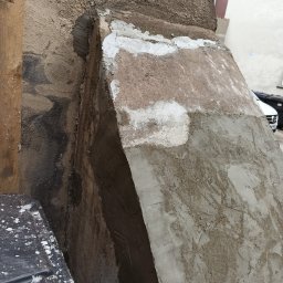 EIT - Narożnik budynku w trakcie prac tynkarskich z widocznymi warstwami zaprawy i częściowo nałożonym tynkiem, drewniana listwa z boku.