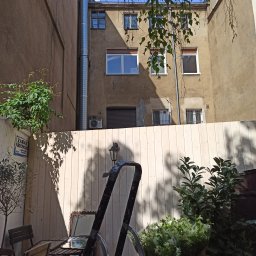 EIT - Nowy, jasny płot drewniany w zacisznym podwórku kamienicy, z widocznymi roślinami i elementami dekoracyjnymi, tworzący przytulną przestrzeń.