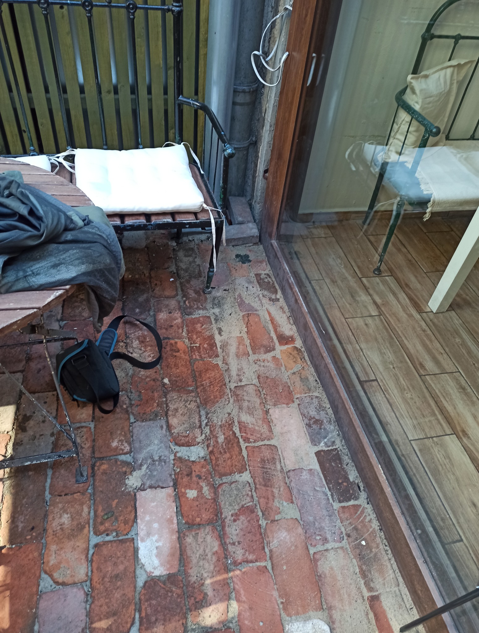 Ceglana posadzka na zewnątrz budynku, z widocznym fragmentem metalowej ławki z poduszką i torbą obok. W tle widoczne wnętrze przez szklane drzwi.