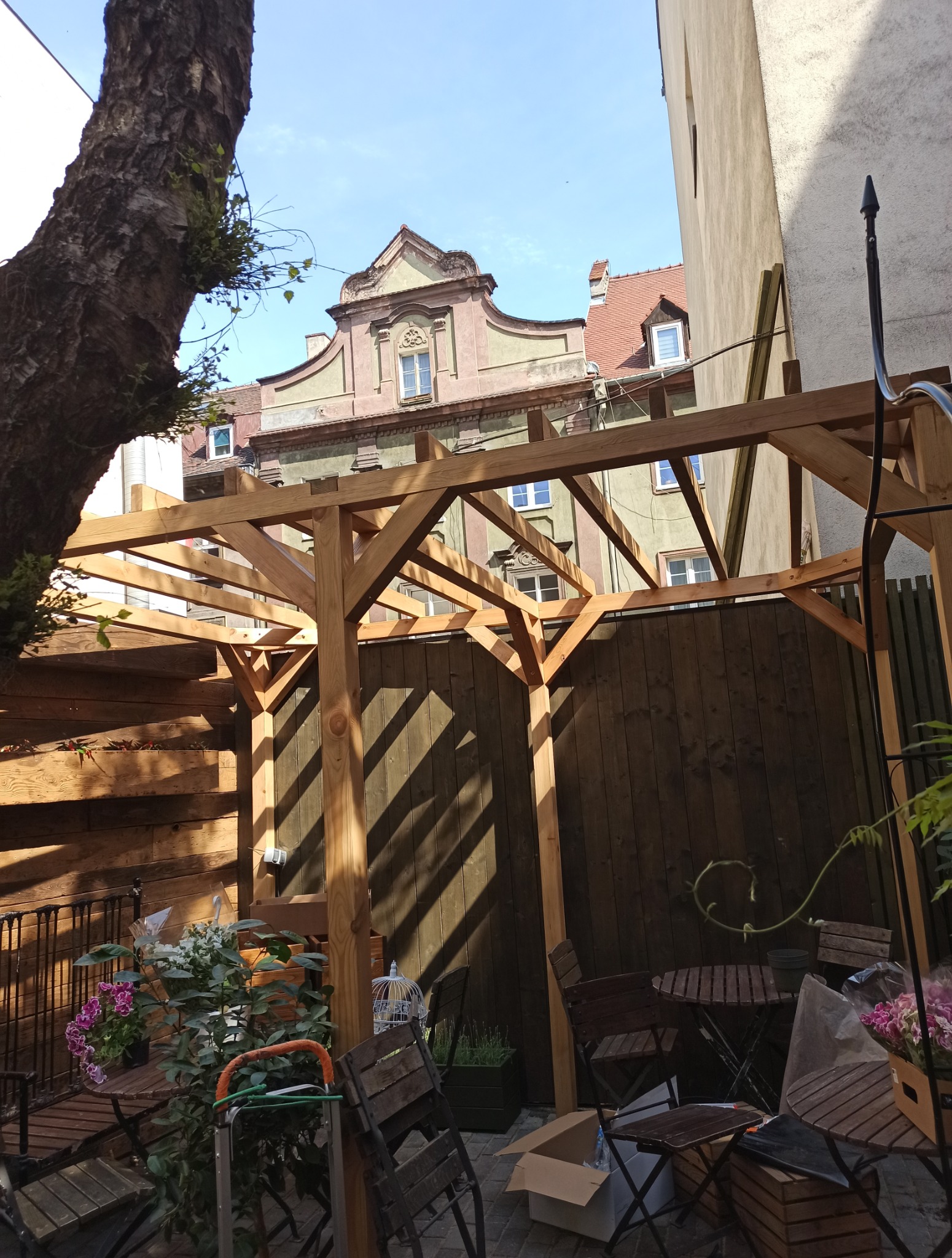 Drewniana konstrukcja pergoli w ogrodzie, widok na kamienicę w tle, fragment drzewa po lewej. Meble ogrodowe i rośliny w otoczeniu. Cień na drewnianym płocie.