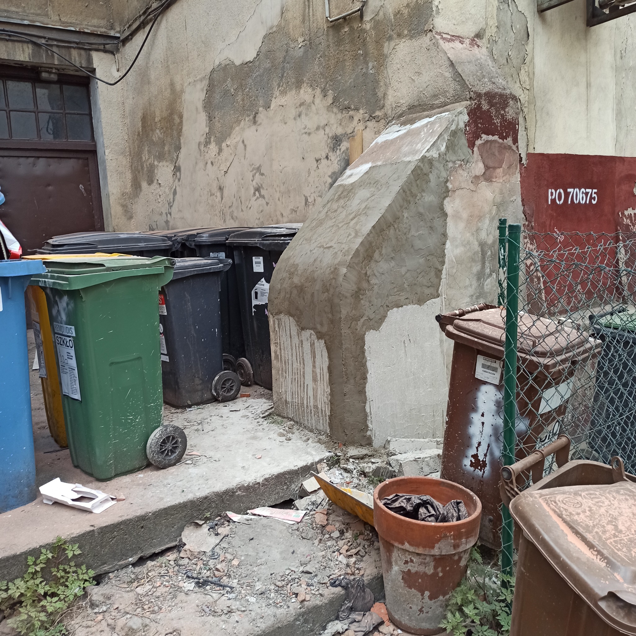 Narożna elewacja budynku w trakcie naprawy, obok śmietniki w różnych kolorach i doniczka. Widoczne ślady uszkodzeń i napraw na ścianie.