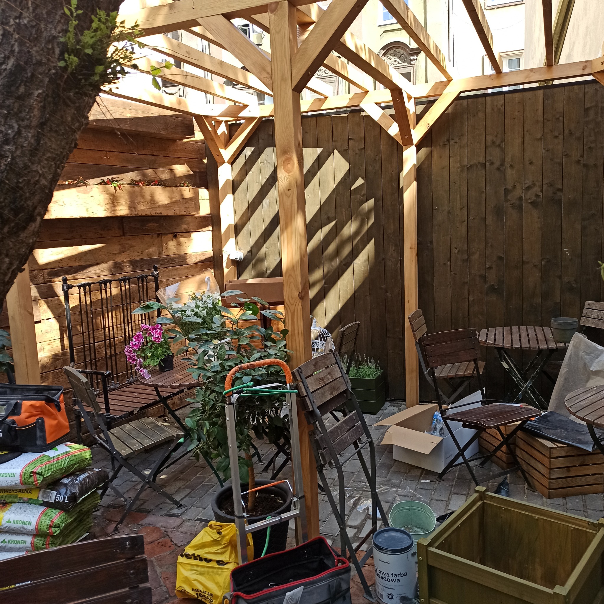 Drewniana pergola w trakcie budowy na brukowanym patio, narzędzia i worki z ziemią w tle, częściowo zacieniona. Widoczne elementy konstrukcyjne i narzędzia pracy.