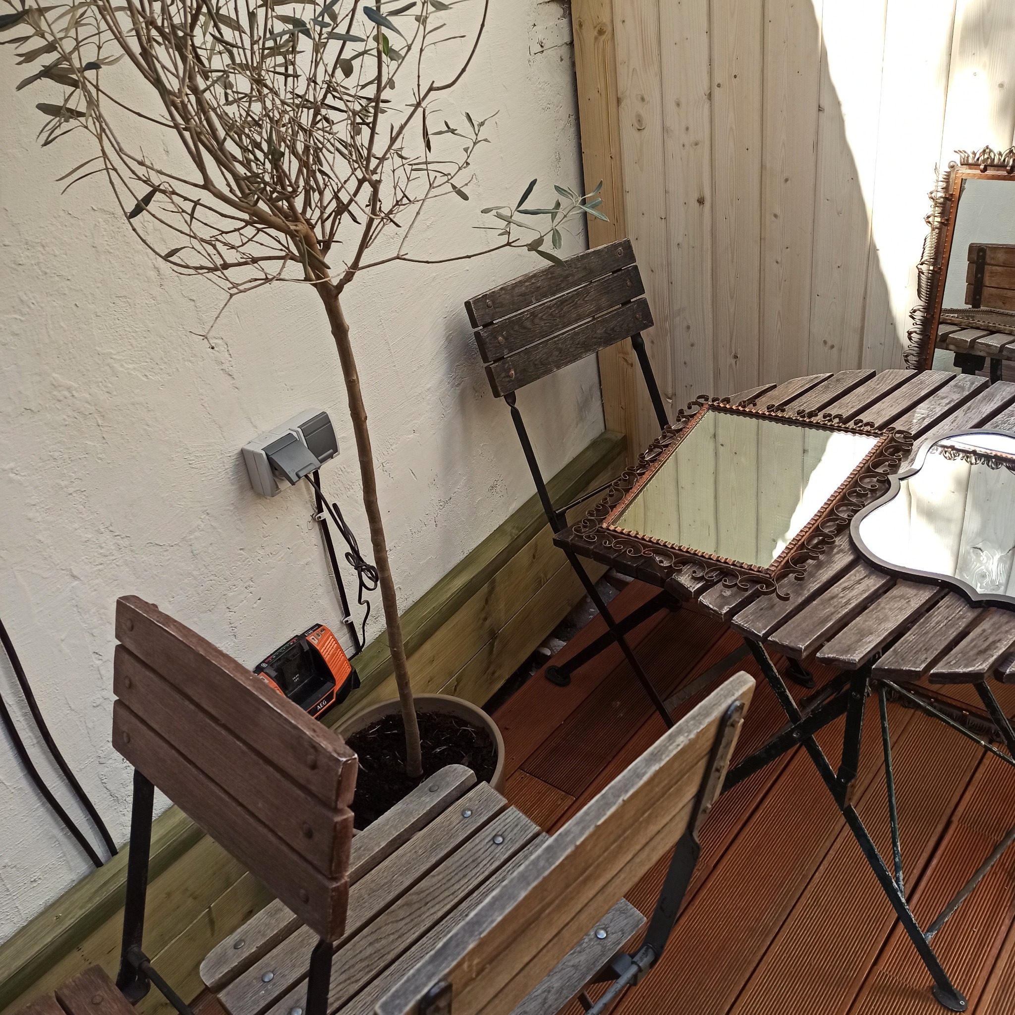 Rustykalny balkon z drewnianą podłogą i meblami. Na stole dwa ozdobne lustra, obok doniczka z drzewkiem. Ściana z gniazdkiem i narzędziem.