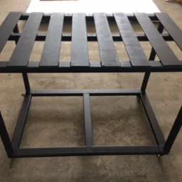 Czarna, metalowa konstrukcja na kółkach, z poziomymi poprzeczkami tworzącymi rodzaj półki. Widoczne spawane łączenia i surowy wygląd metalu.