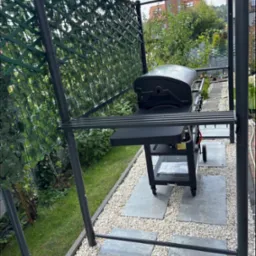 Zadaszona altana z grillem na kamiennej ścieżce, otoczona zielenią. Nowoczesny design,  elementy dekoracyjne z siatki i roślin.  Widok na zadbany ogród i dom w tle.