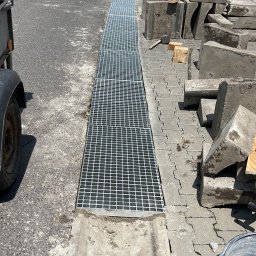 Krzysztof Dominik - Ułożona krata odpływowa z betonu na kostce brukowej. Obok leżą betonowe elementy i narzędzia. Widok z góry na ulicę i fragment budynku.