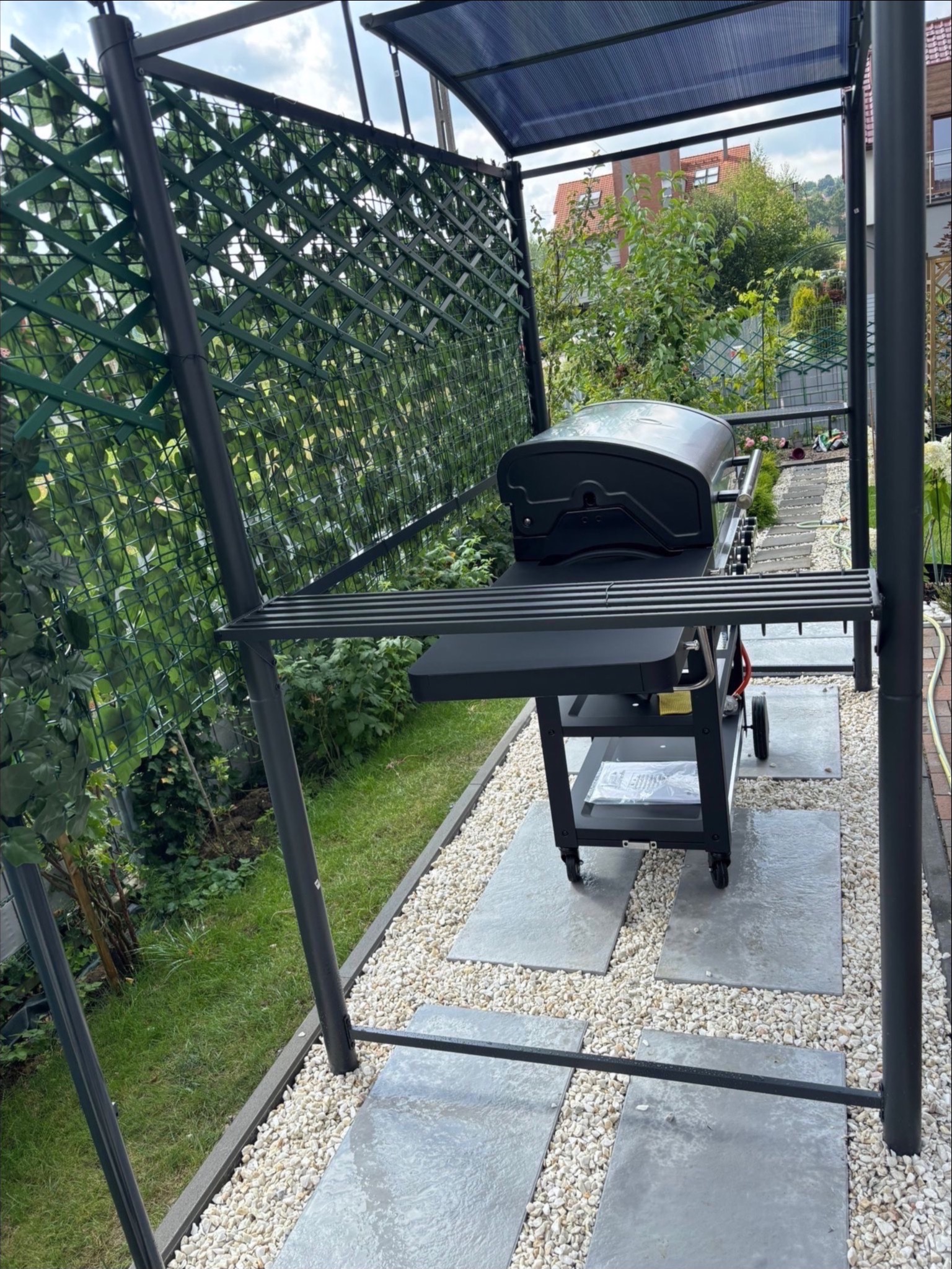 Zadaszona altana z grillem na kamiennej ścieżce, otoczona zielenią. Nowoczesny design,  elementy dekoracyjne z siatki i roślin.  Widok na zadbany ogród i dom w tle.