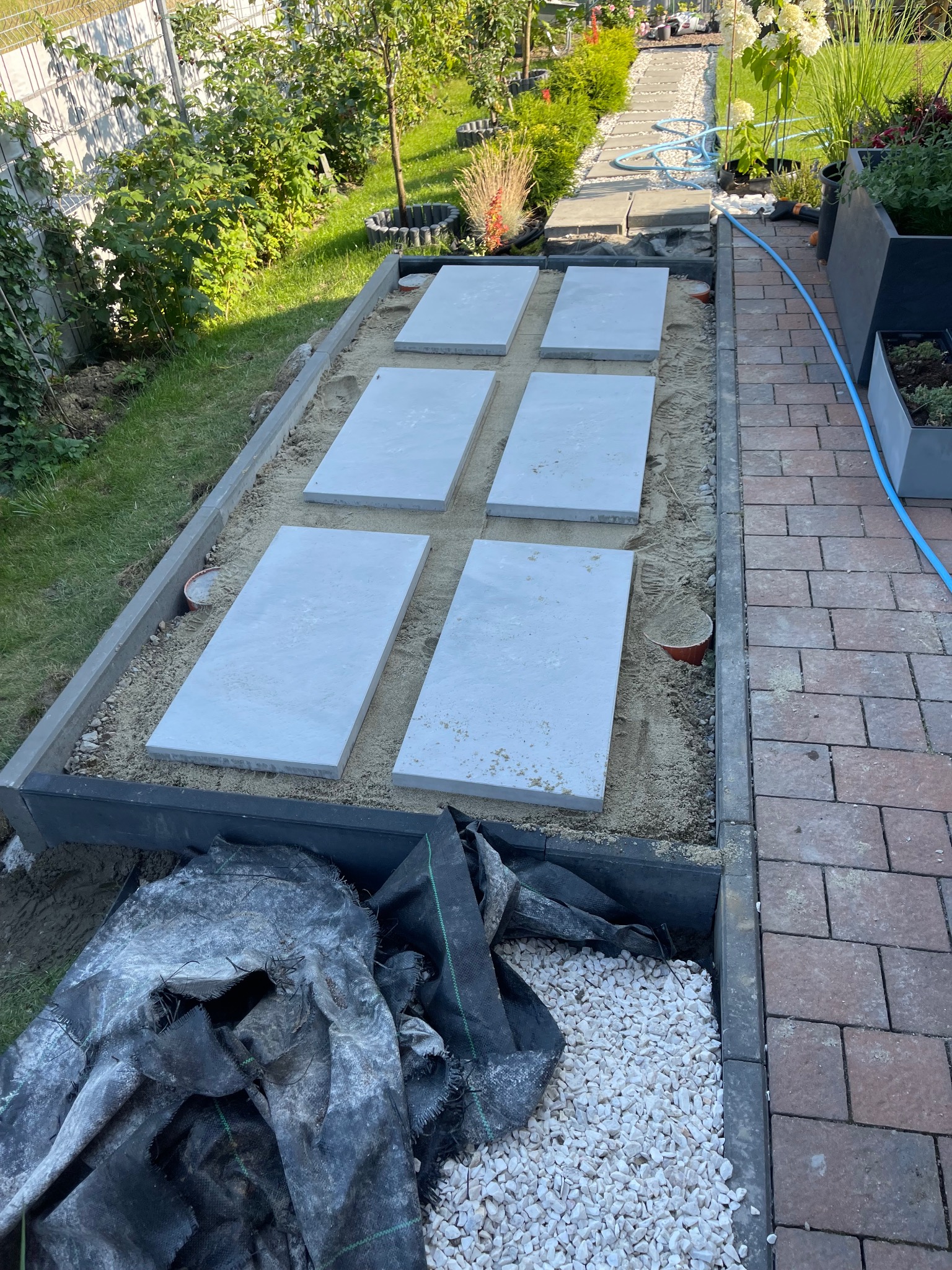 Montaż ścieżki z płyt betonowych w ogrodzie, widoczne etapy prac ziemnych i podsypka piaskowa, obrzeża z kostki, w tle trawnik i roślinność.