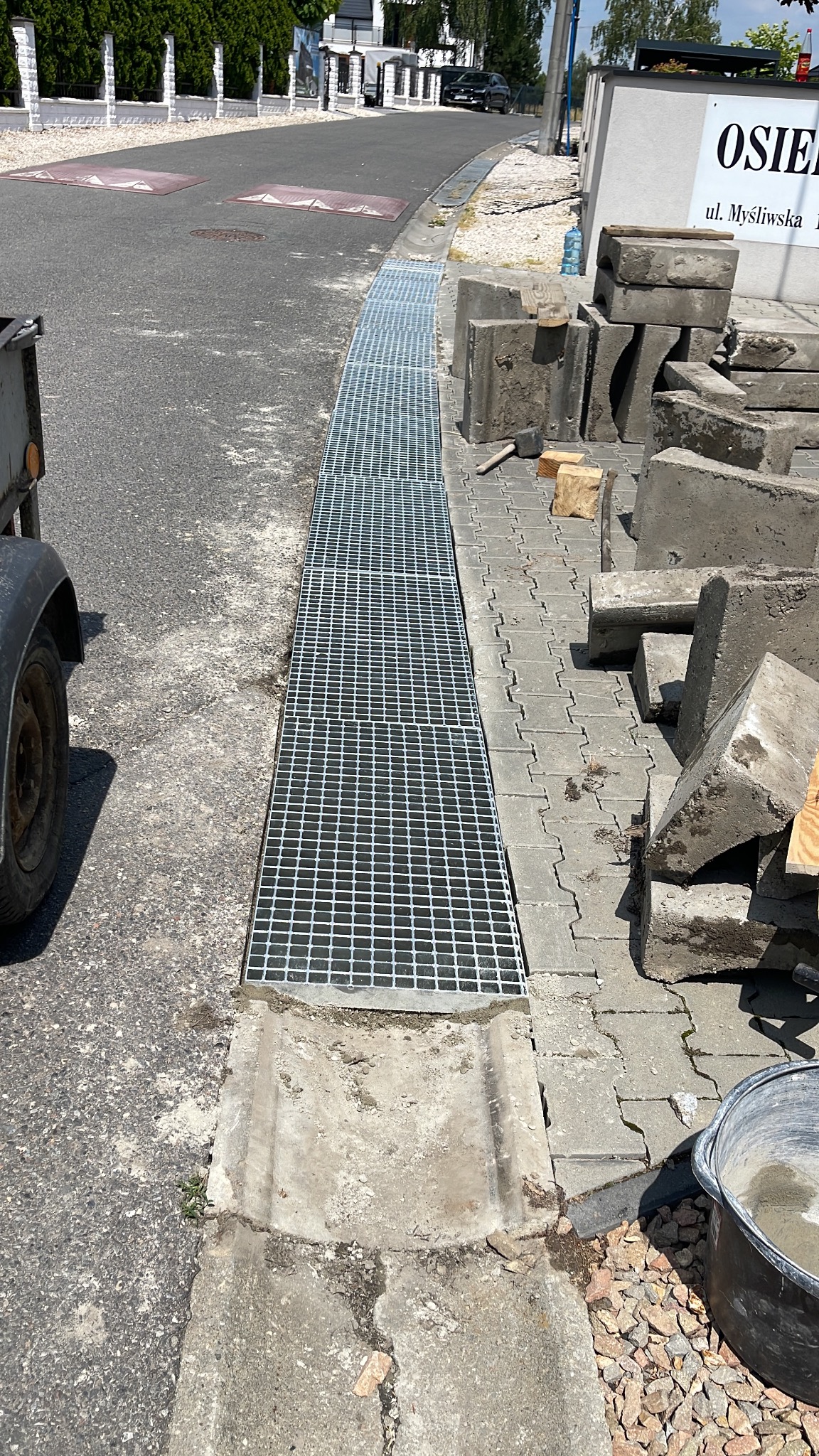 Ułożona krata odpływowa z betonu na kostce brukowej. Obok leżą betonowe elementy i narzędzia. Widok z góry na ulicę i fragment budynku.
