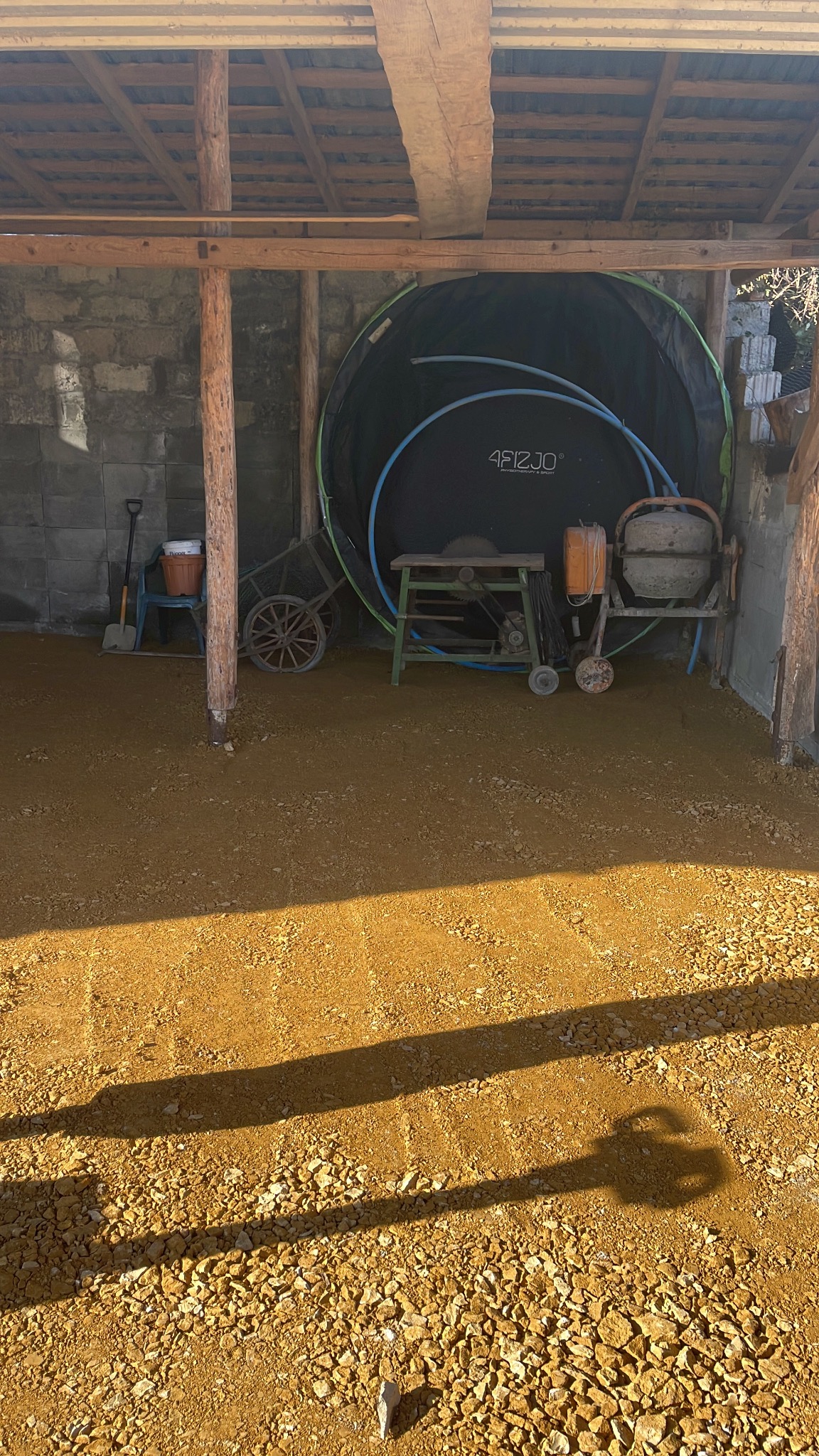 Przygotowanie podłoża pod altanę: wyrównany teren z drobnym kruszywem, cień konstrukcji, narzędzia budowlane w tle, w tym betoniarka. Widoczne bloczki betonowe.