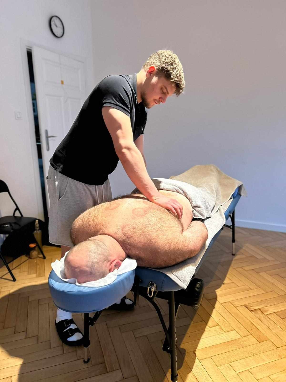 Mężczyzna w czarnej koszulce wykonuje masaż pleców pacjenta leżącego na niebieskim stole do masażu. Widoczne ślady po bańkach.