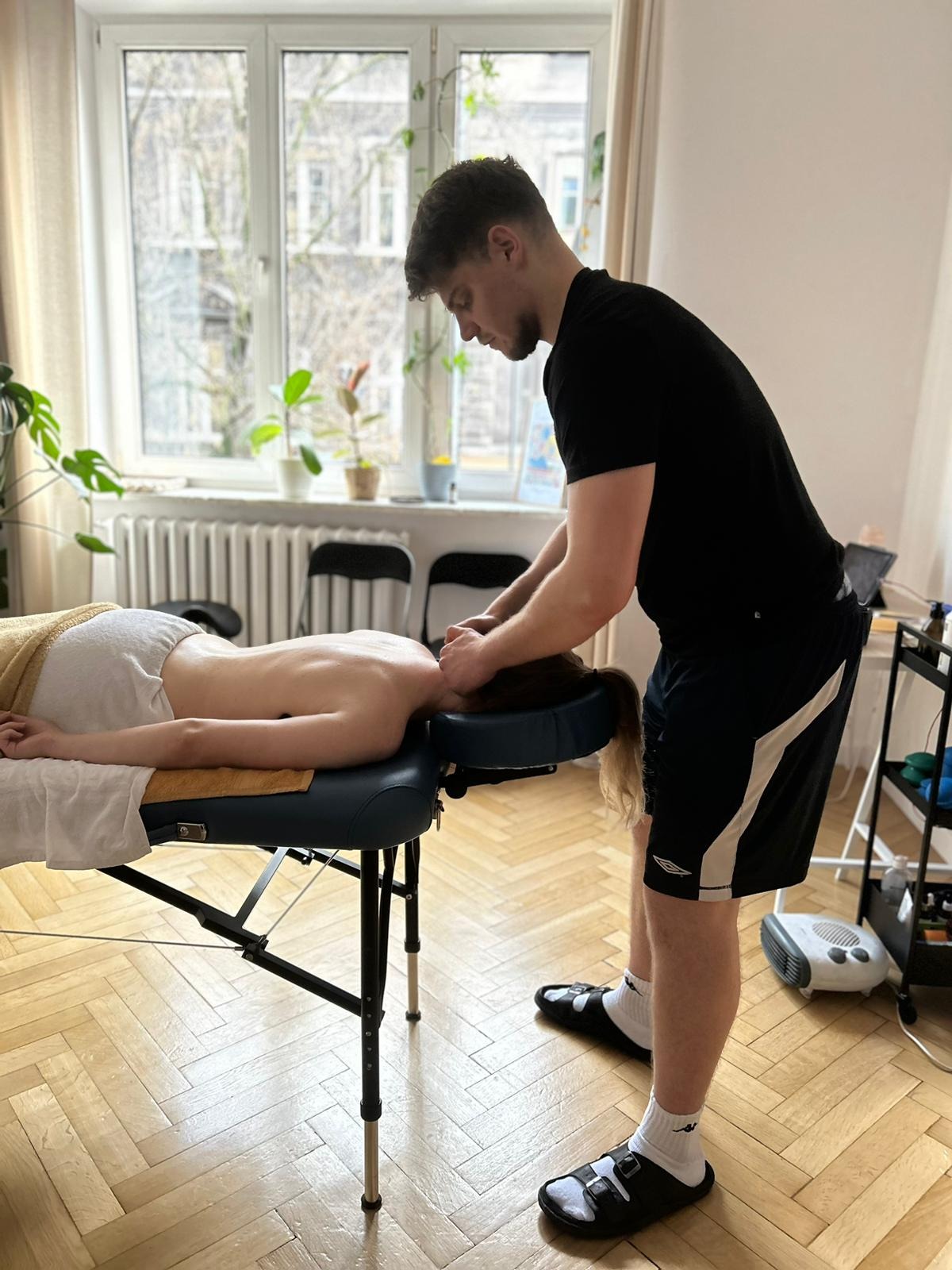 Mężczyzna wykonuje masaż głowy kobiecie leżącej na niebieskim stole do masażu. W tle okno z widokiem na budynek i rośliny doniczkowe. Parkiet w jodełkę.