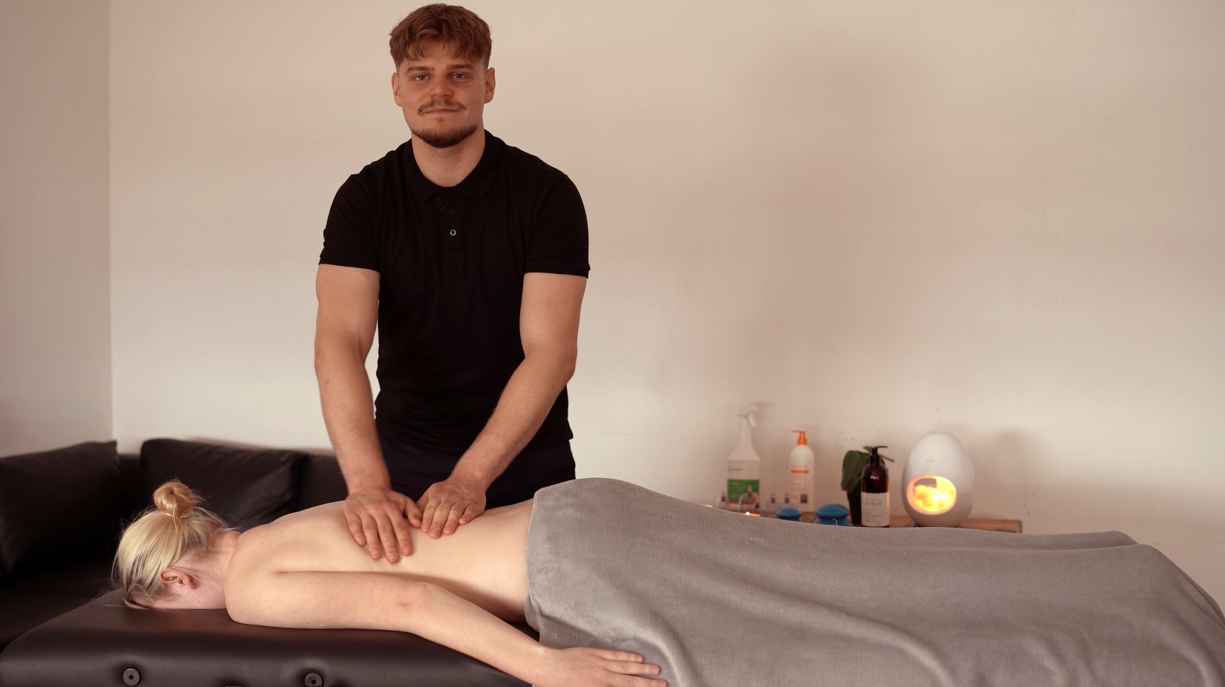 Masażysta w czarnej koszulce wykonuje masaż pleców klientki leżącej na stole do masażu, relaksująca atmosfera, lampka w tle, neutralne kolory.