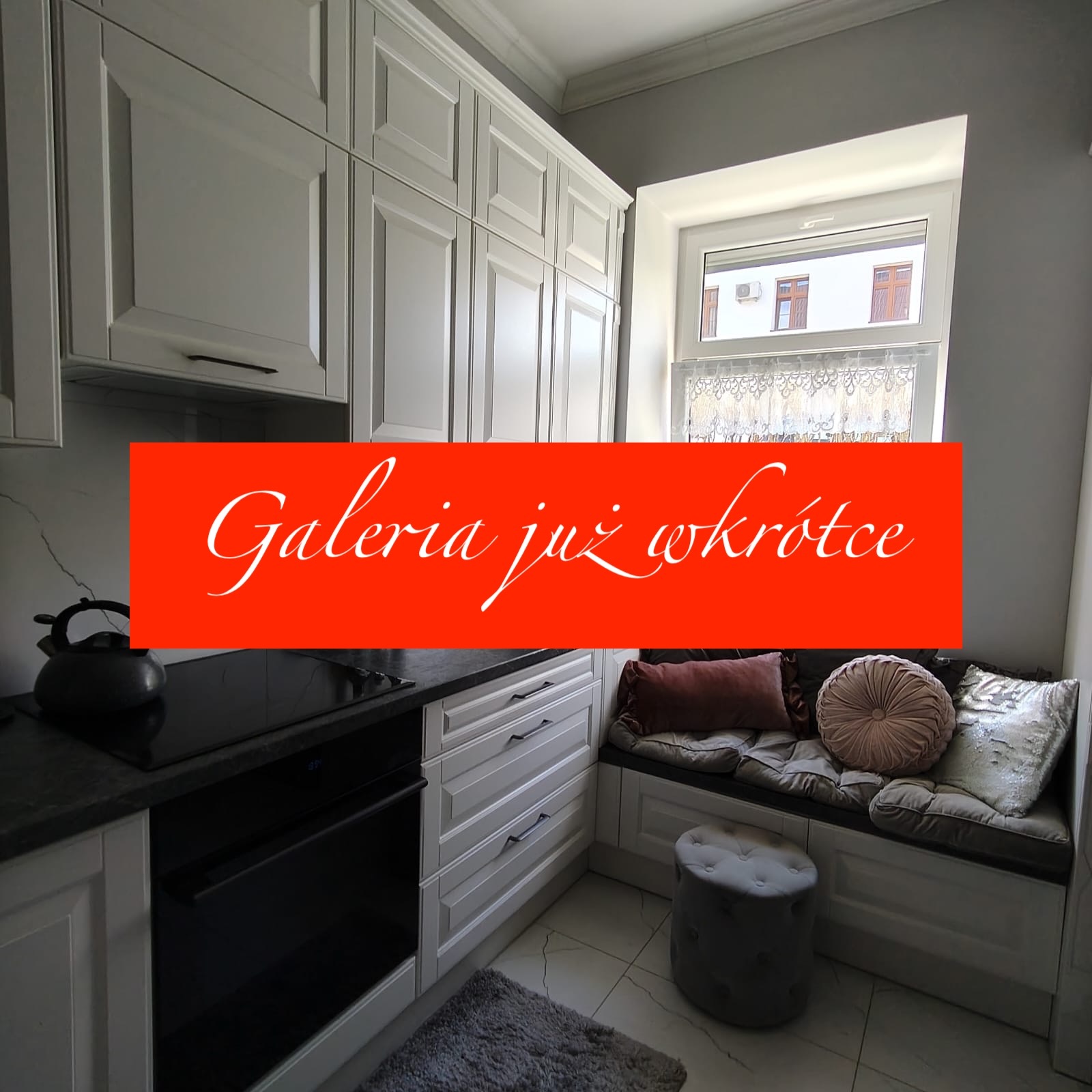 Jasna kuchnia w stylu klasycznym z zabudową pod oknem. Białe fronty szafek, ciemny blat i sprzęt AGD. W oknie widok na budynek.