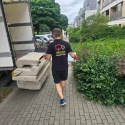 Pracownik w czarnej koszulce z logo firmy transportowej przenosi jasne sofy z busa na chodnik z kostki brukowej. W tle zieleń i budynki mieszkalne.
