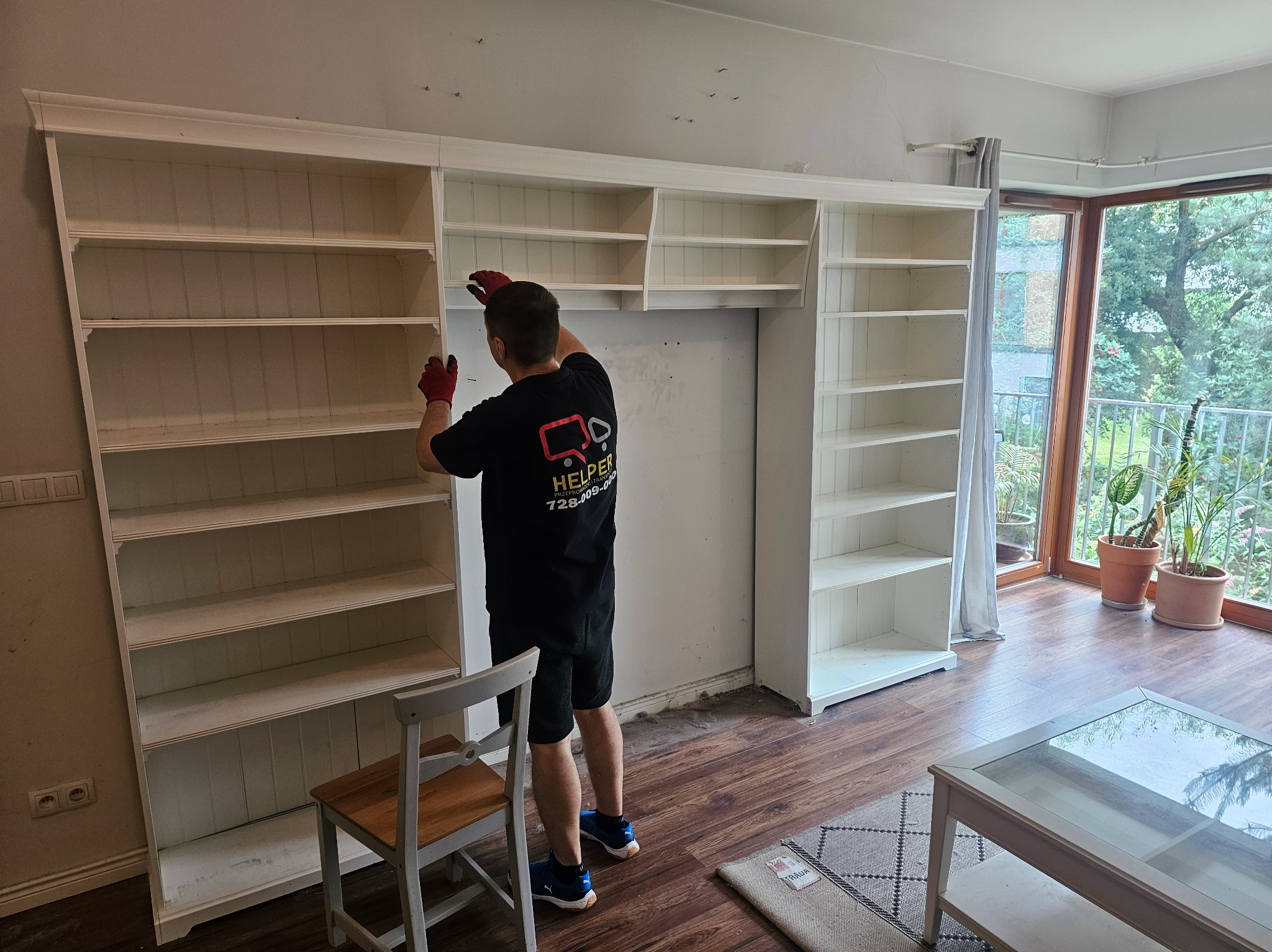 Pracownik w czerwonych rękawicach montuje biały regał z półkami w jasnym pokoju z dużym oknem balkonowym. Widoczny fragment krzesła i stolika.