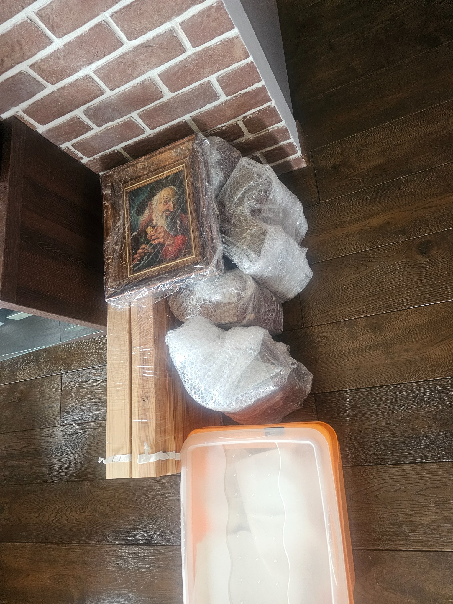 Obraz w ramie, drewniane elementy i karton zapakowane w folię bąbelkową, przygotowane do transportu na brązowej podłodze. Widoczna ściana z cegły.