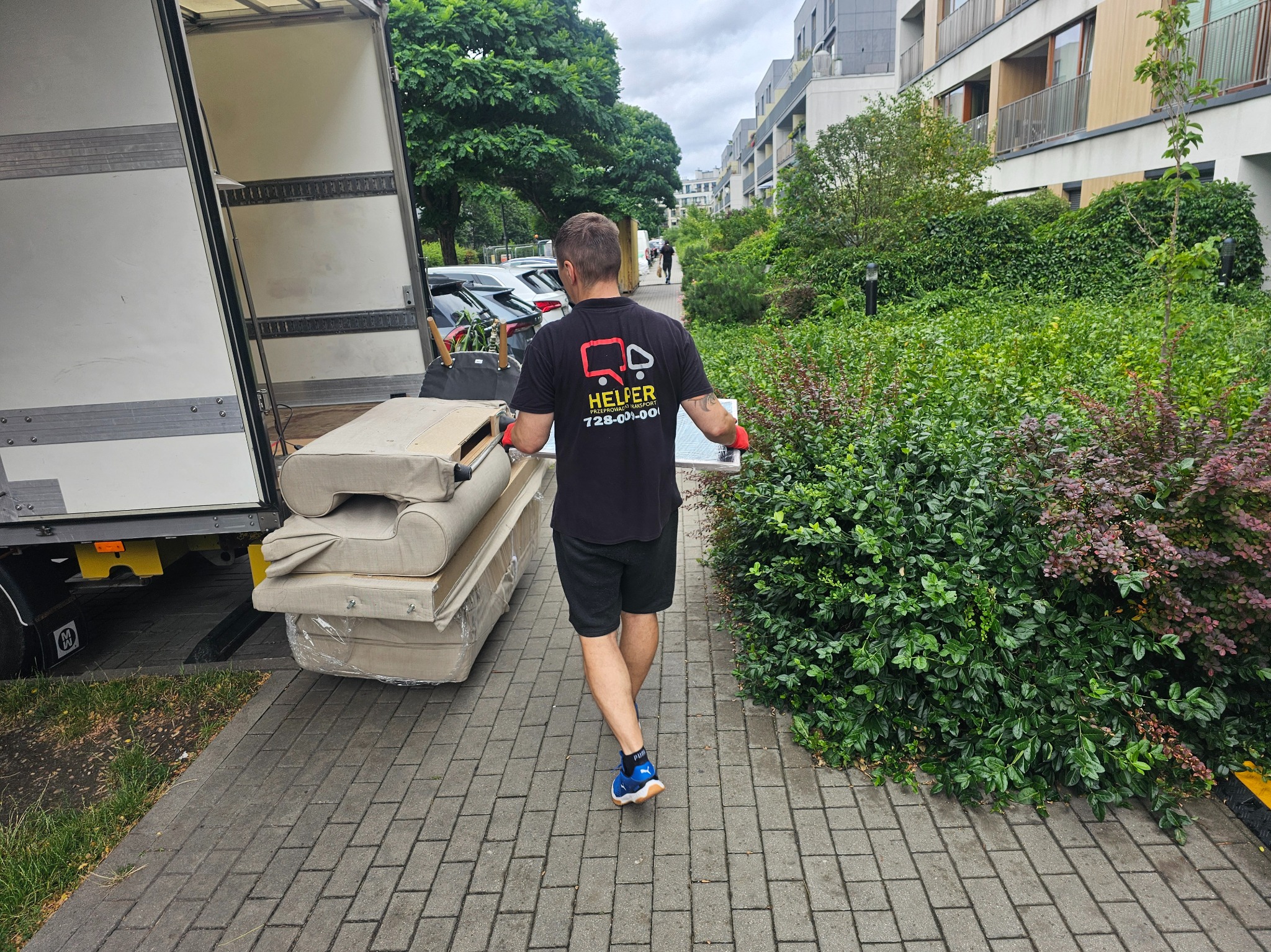 Pracownik w czarnej koszulce z logo firmy transportowej przenosi jasne sofy z busa na chodnik z kostki brukowej. W tle zieleń i budynki mieszkalne.