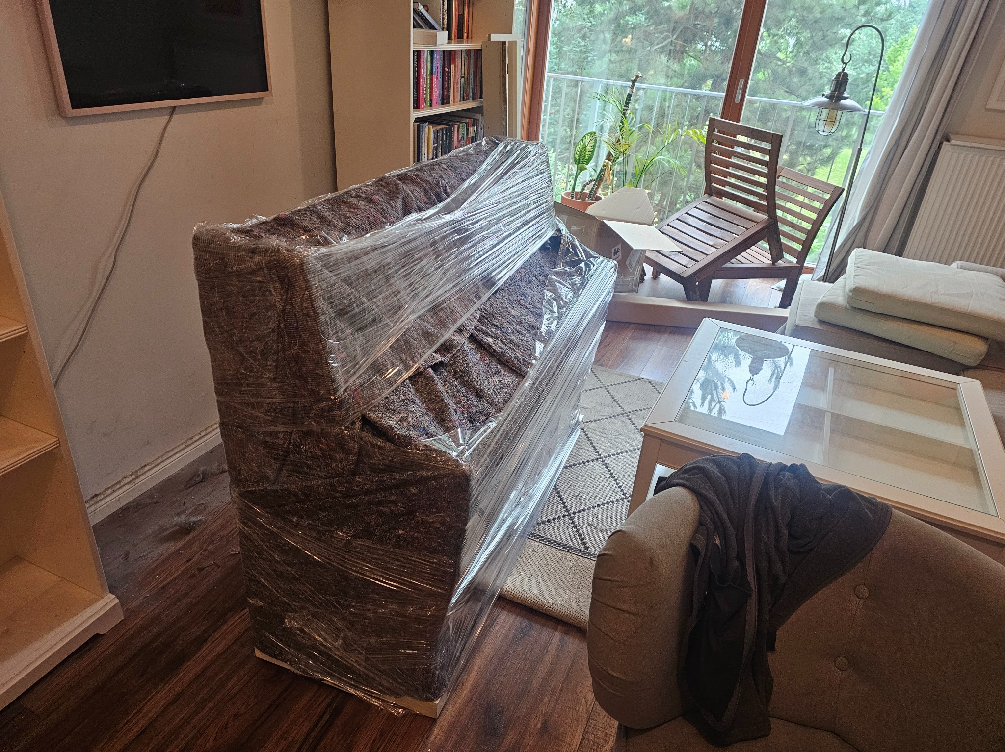 Pianino zabezpieczone folią stretch, gotowe do transportu w Piasecznie. Widoczne meble w tle sugerują przeprowadzkę. Ochrona przed uszkodzeniami.