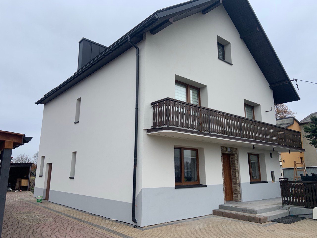 Dwukondygnacyjny dom z balkonem po renowacji elewacji, z białymi ścianami i szarym cokołem. Widoczne brązowe okna i balustrada balkonu, czarne rynny i dach.