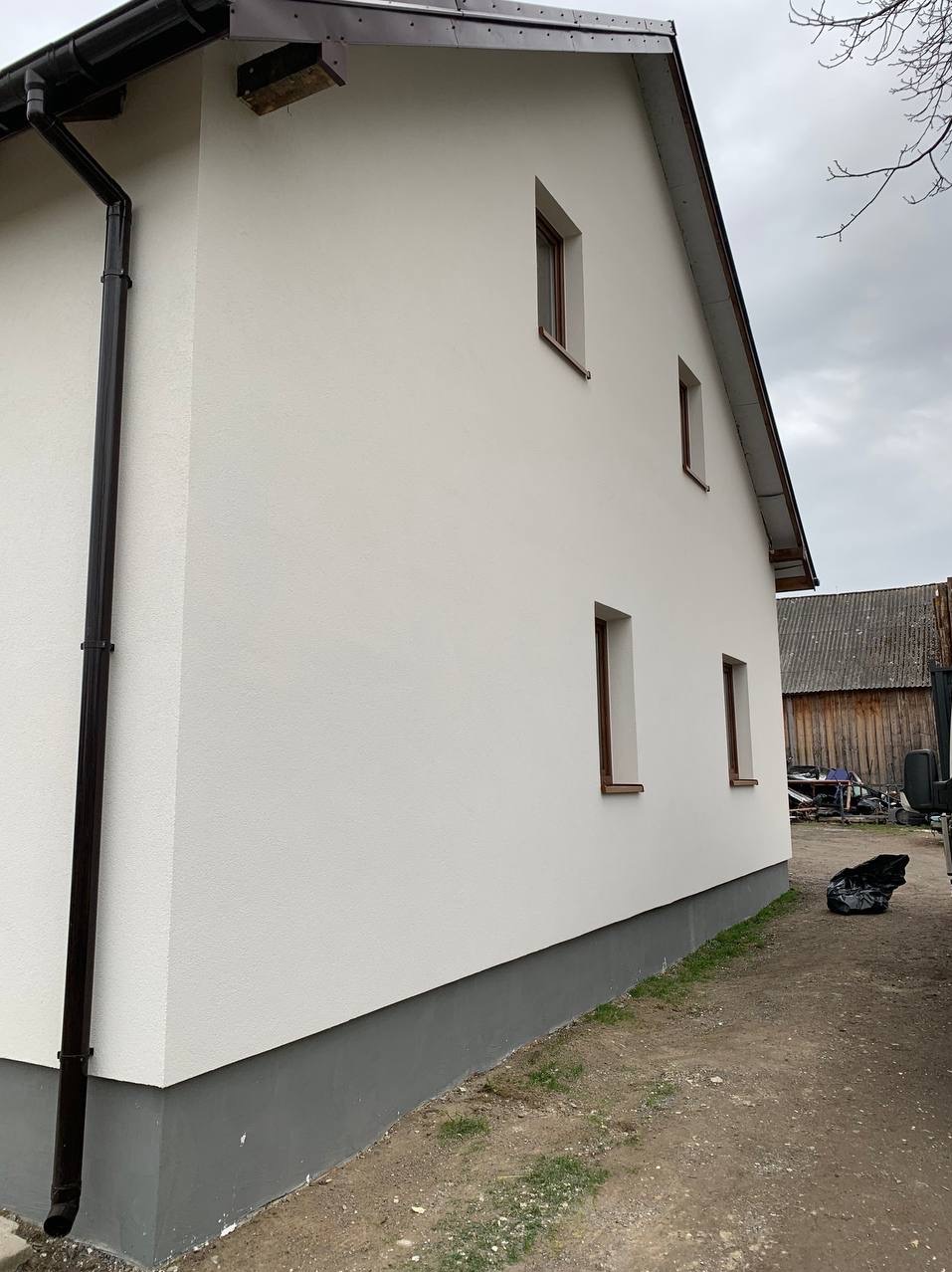 Boczny widok domu po renowacji elewacji: biała ściana z brązowymi oknami, ciemny drenaż, szara podmurówka i widoczne tło z budynkiem gospodarczym.