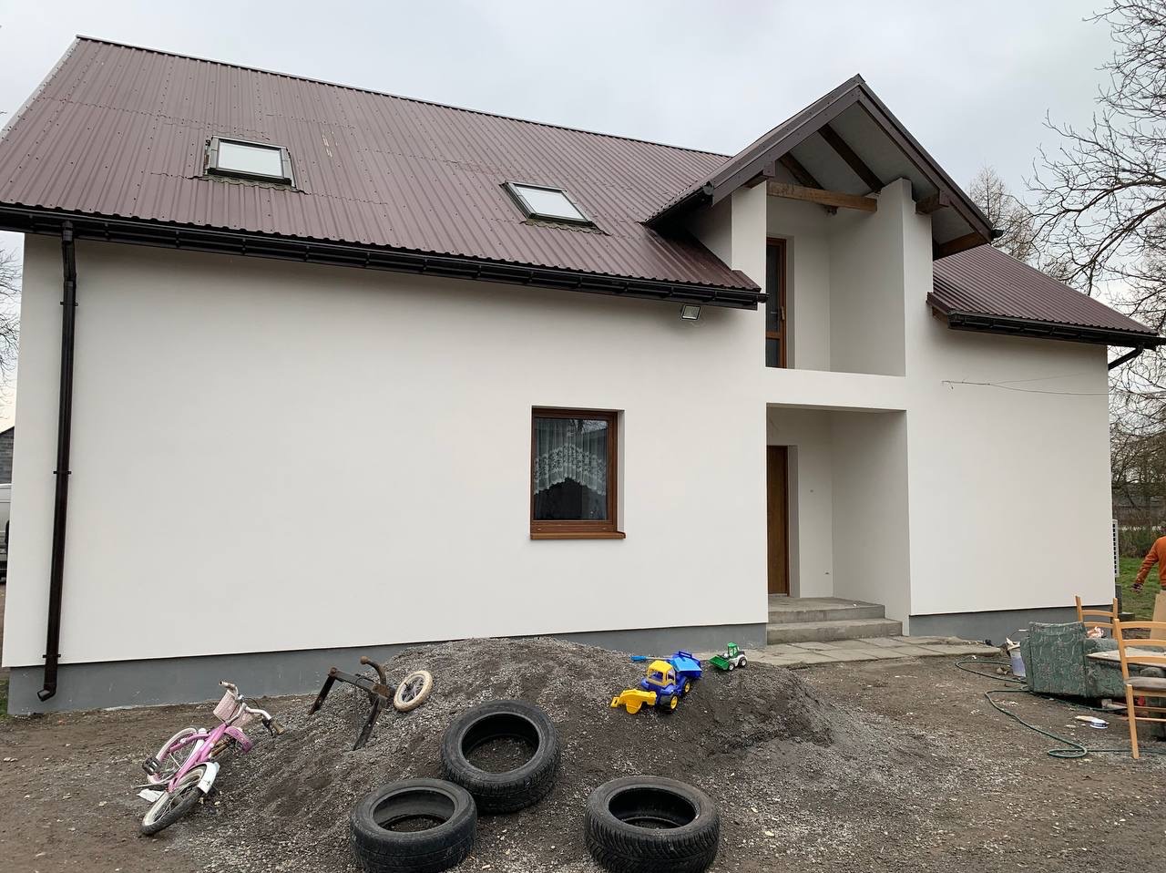 Biała elewacja domu jednorodzinnego z brązowym dachem, widok z poziomu gruntu, zabałaganione podwórko z oponami i zabawkami. Nowoczesna architektura.