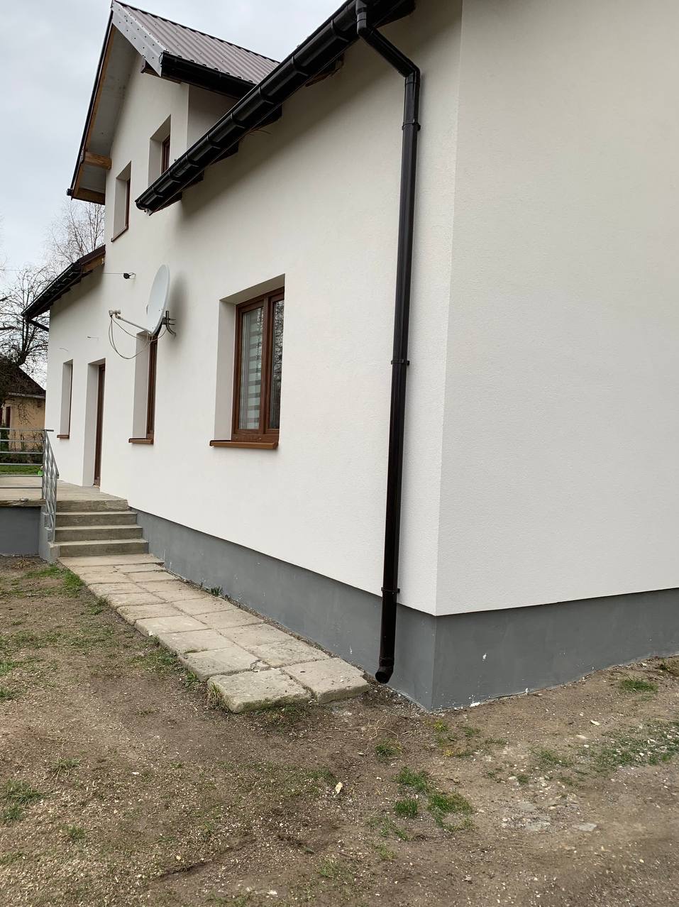 Boczny widok domu po renowacji elewacji w Ozorkowie. Biała ściana z brązowymi oknami i rynnami. Widoczne schody i chodnik z płyt.