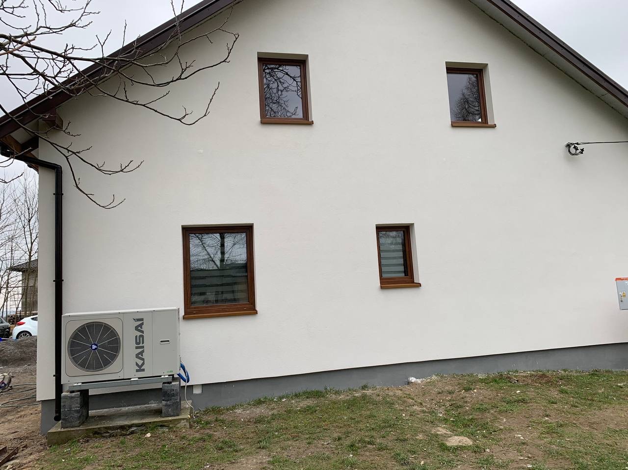Biała elewacja domu z brązowymi oknami i zamontowaną pompą ciepła Kaisai na zewnątrz. Widoczne drzewa bez liści i trawa. Dom po ociepleniu.