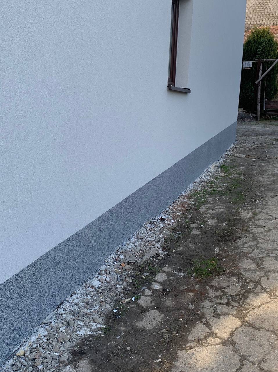 Ściana budynku po tynkowaniu z widocznym cokołem w szarym kolorze. Nowa elewacja w jasnym odcieniu, okno z brązową ramą. Widok z zewnątrz na fragment budynku.