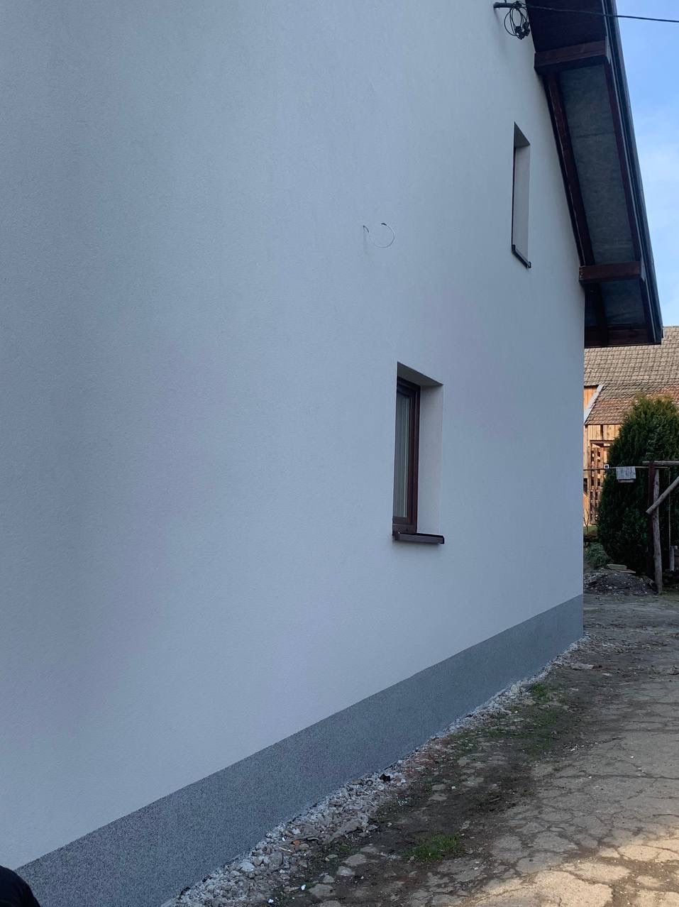 Świeżo otynkowana, biała ściana domu z dwoma oknami o brązowych ramach. Dolna część ściany wykończona szarym tynkiem. Widok z boku, na tle zieleni i fragmentu dachu sąsiedniego budynku.