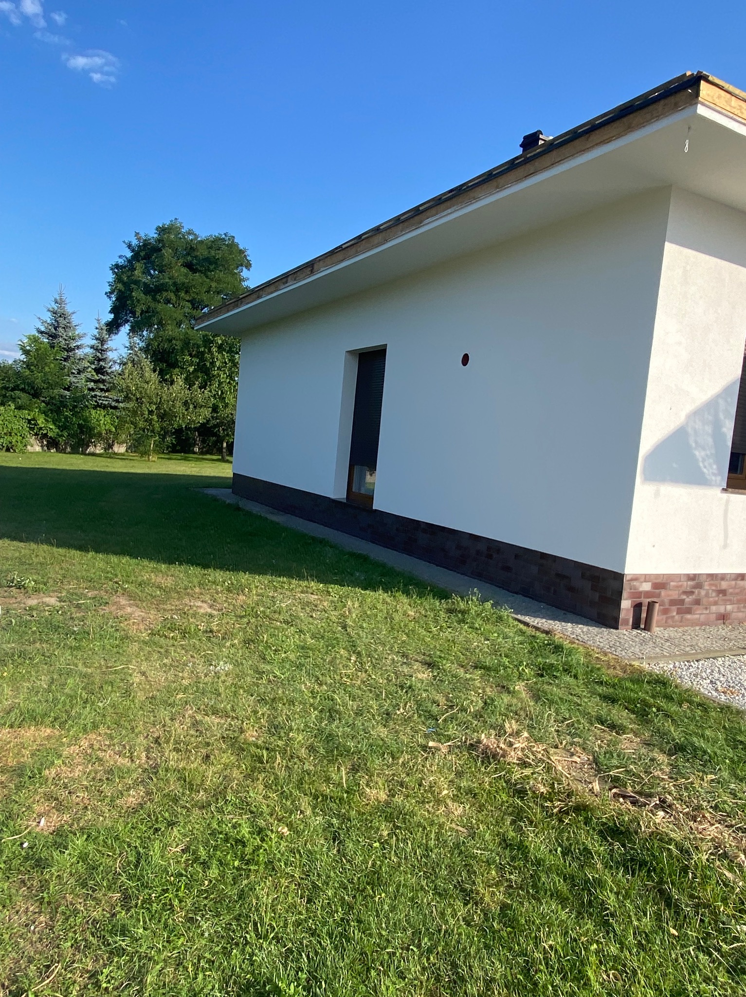 Biała elewacja parterowego domu z ciemną podmurówką i widocznym fragmentem dachu, na tle zielonego trawnika i drzew w słoneczny dzień.