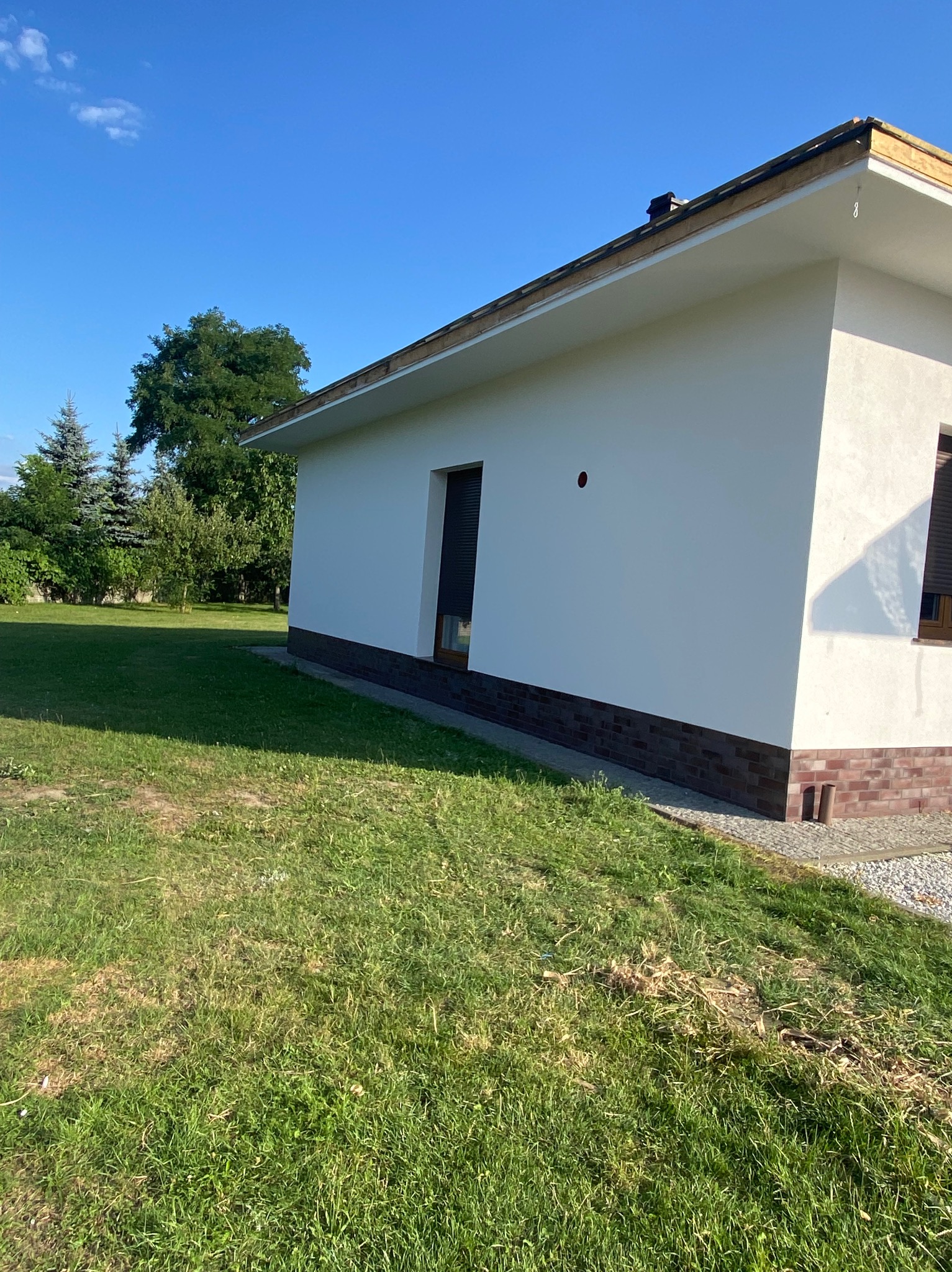 Boczny widok jednopiętrowego domu z białą elewacją i ceglaną podmurówką, okno z żaluzjami, trawnik i drzewa w tle, słoneczny dzień.