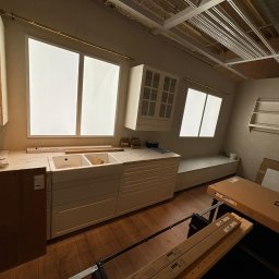 Montaż mebli Jan Borsuk - Wnętrze w trakcie prac: biała zabudowa kuchenna z marmurowym blatem, widoczne okna i elementy konstrukcyjne sufitu. Na podłodze kartony i narzędzia.