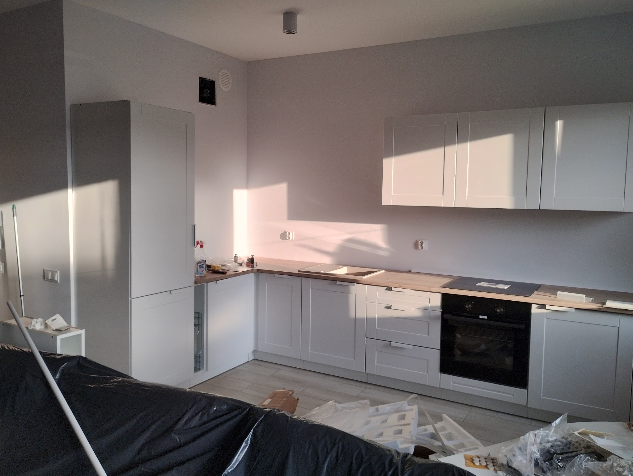 Jasna kuchnia z białymi frontami, drewnianym blatem i czarną płytą grzewczą. Widoczne elementy montażowe i folia ochronna, sugerujące świeży montaż.