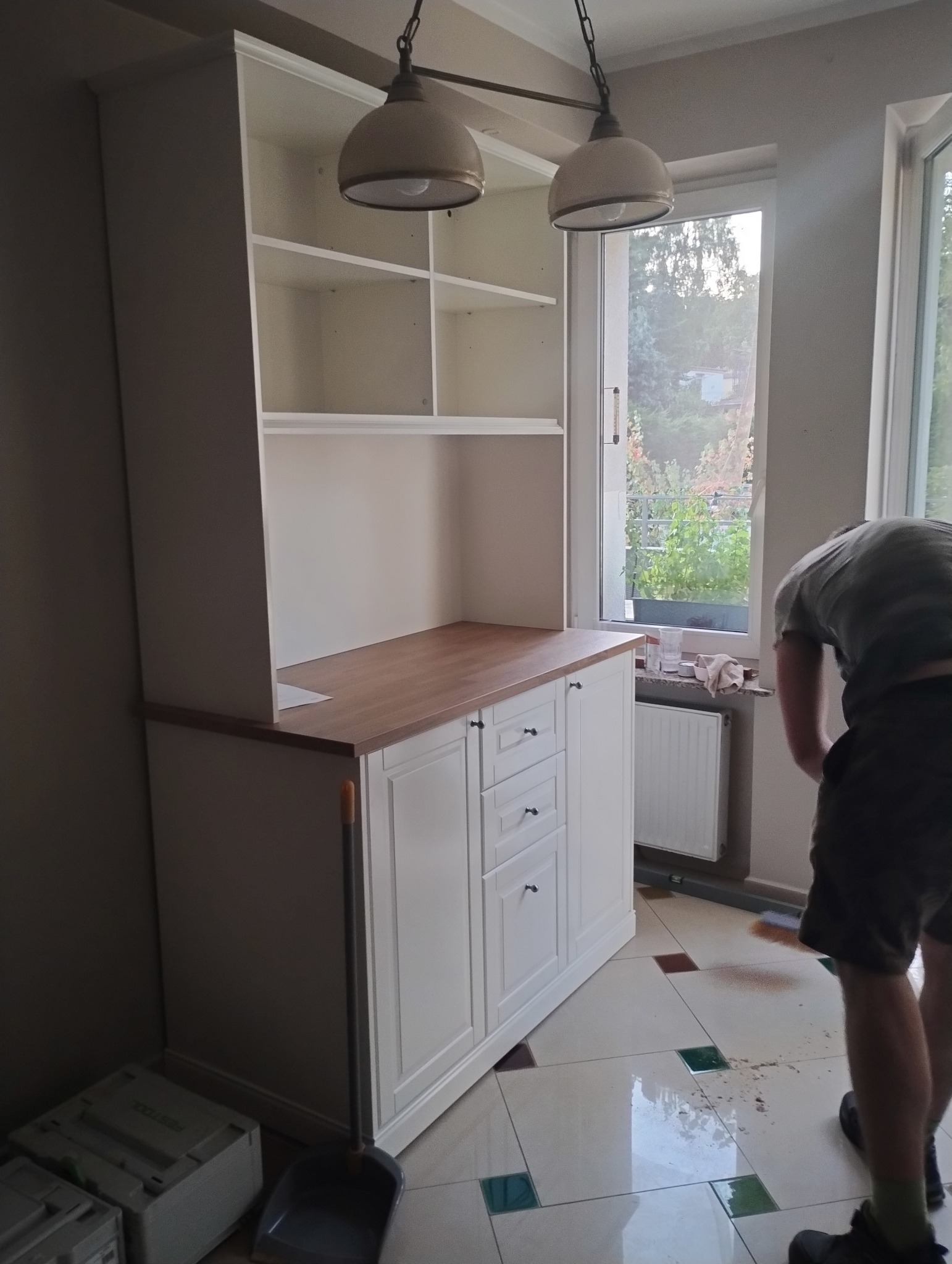 Biała, klasyczna zabudowa kuchenna z drewnianym blatem, częściowo widoczna osoba sprzątająca. Uwagę przykuwają lampy w stylu retro i okno z widokiem na zieleń.