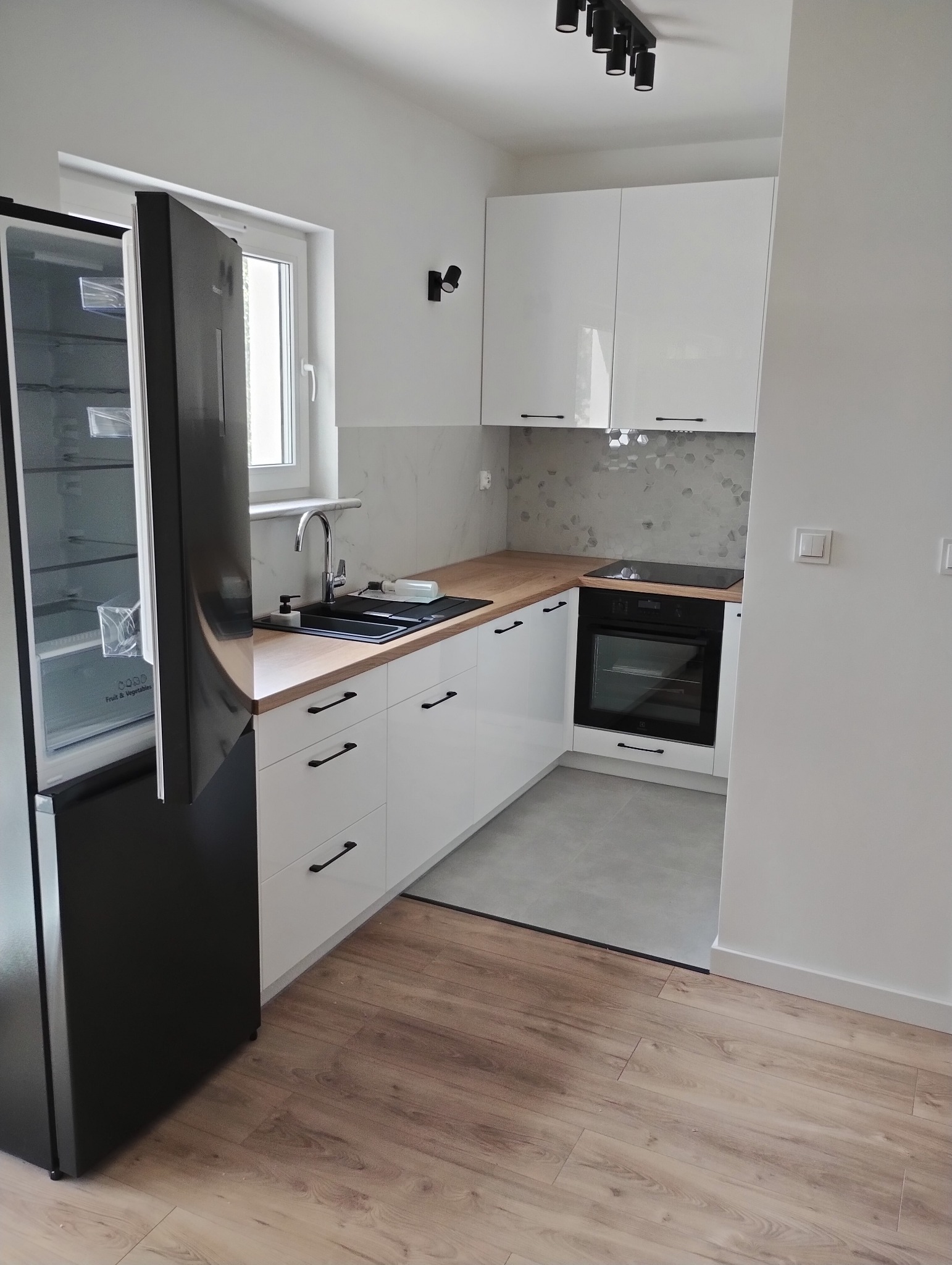 Nowoczesna, jasna kuchnia z białymi szafkami, drewnianym blatem i czarną lodówką z otwartymi drzwiami. Widoczne detale: heksagonalne płytki, czarne uchwyty.