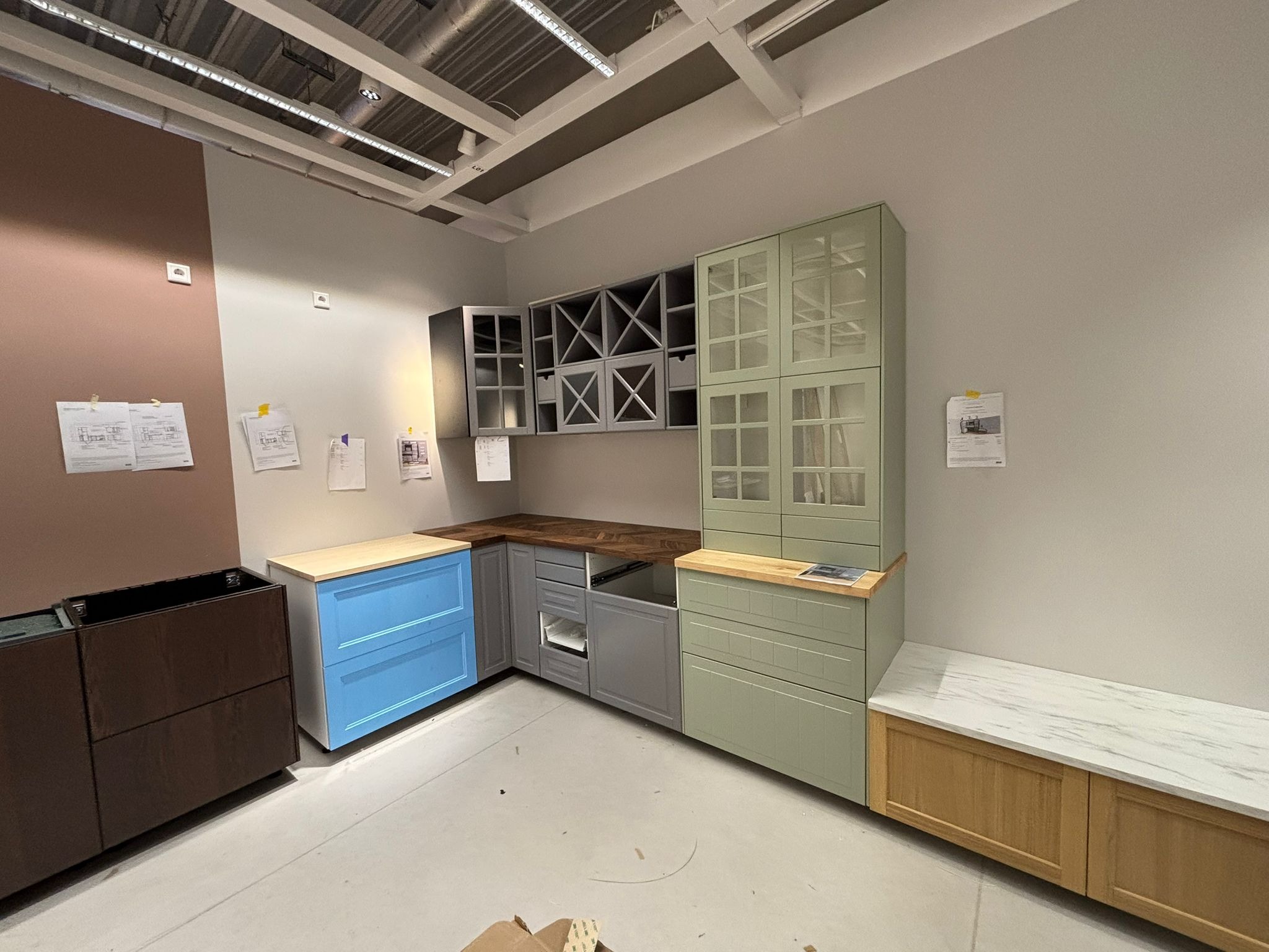 Komplet mebli kuchennych w różnych kolorach: niebieski, szary, zielony i brązowy. Szafki wiszące z przeszklonymi frontami i otwartymi półkami na wino. Blat drewniany.