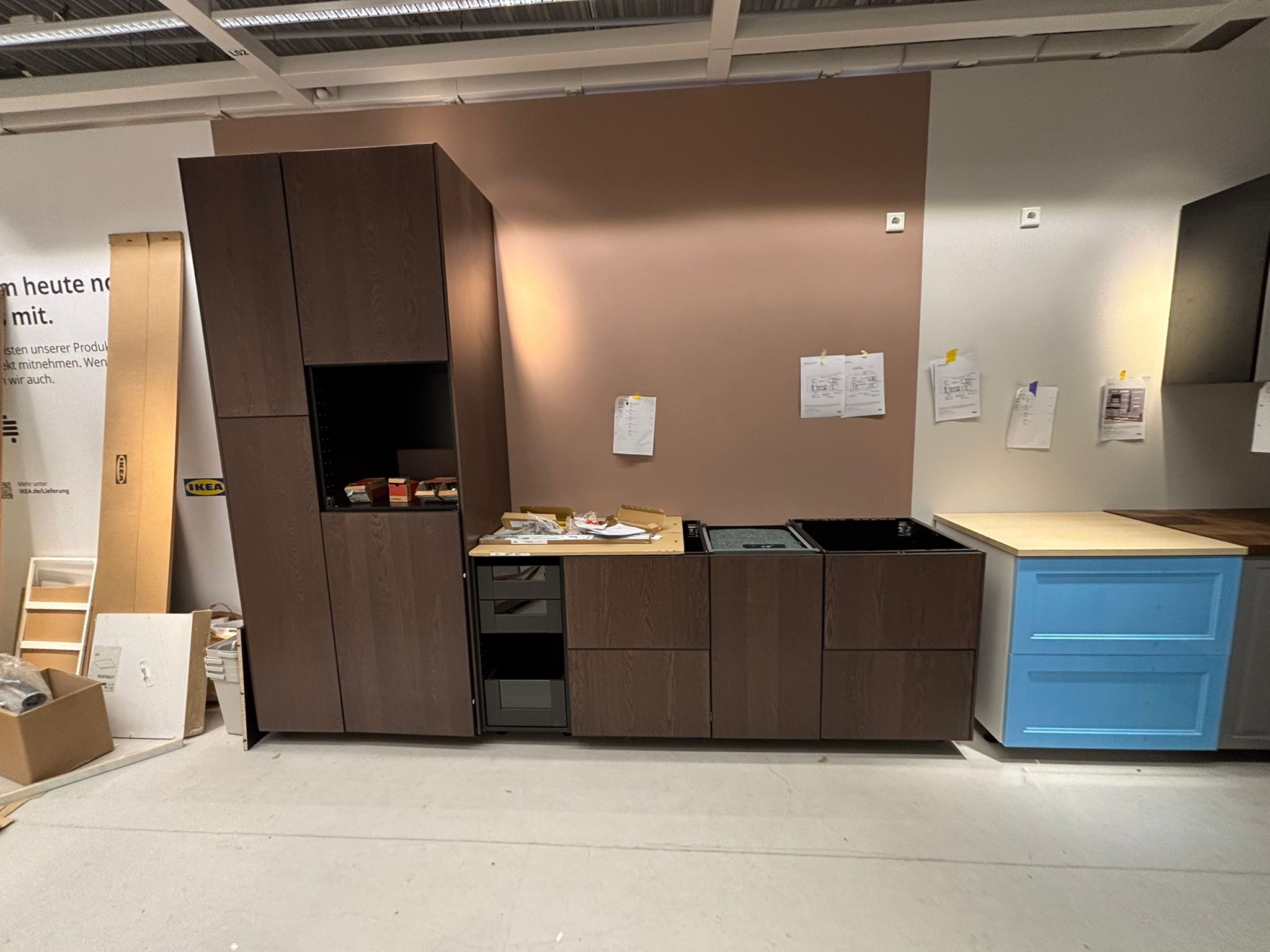 Częściowo zmontowana, nowoczesna kuchnia z ciemnego drewna i niebieskimi akcentami, widoczne kartony i elementy montażowe, sugerujące etap składania.