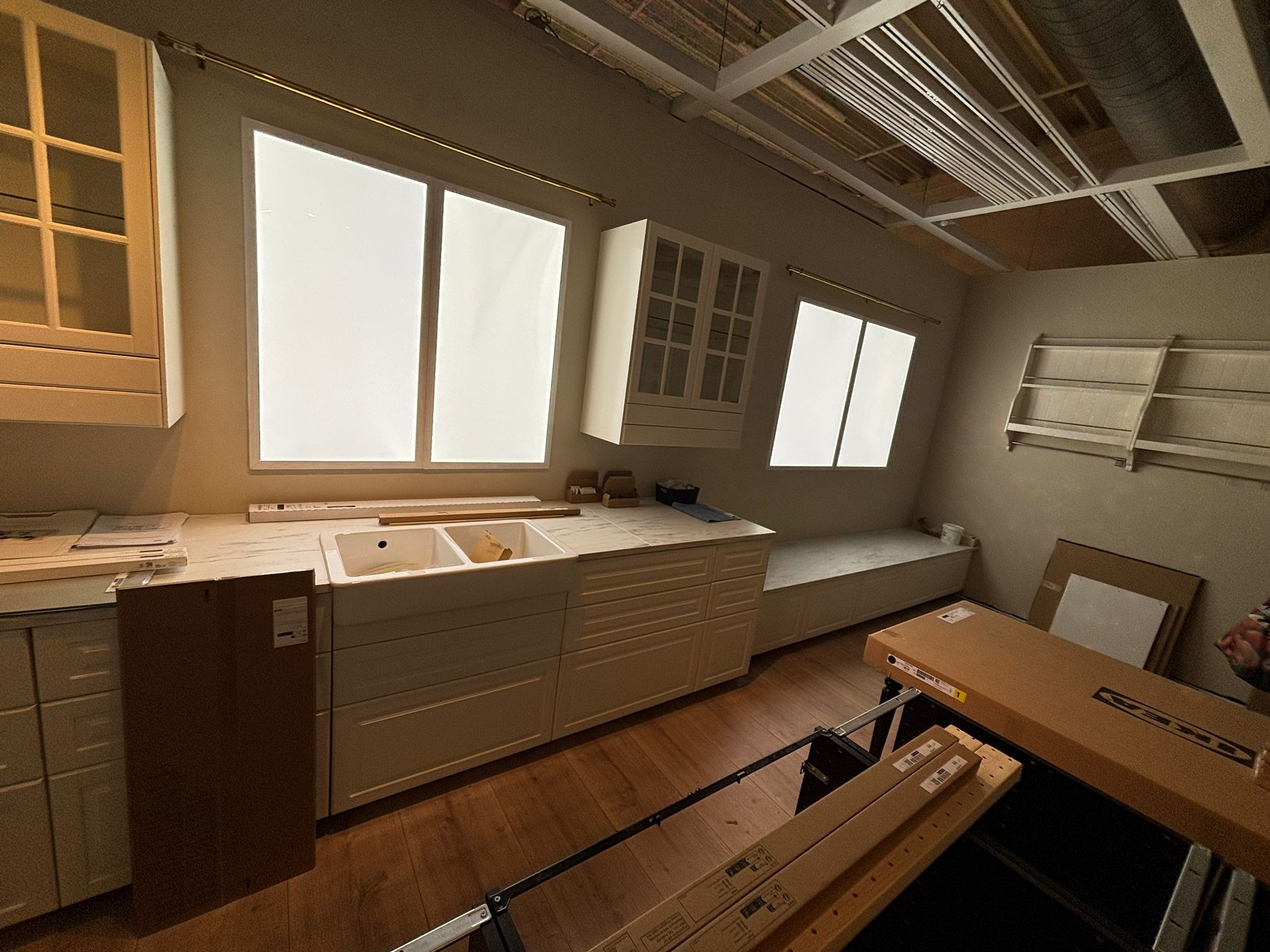 Wnętrze w trakcie prac: biała zabudowa kuchenna z marmurowym blatem, widoczne okna i elementy konstrukcyjne sufitu. Na podłodze kartony i narzędzia.